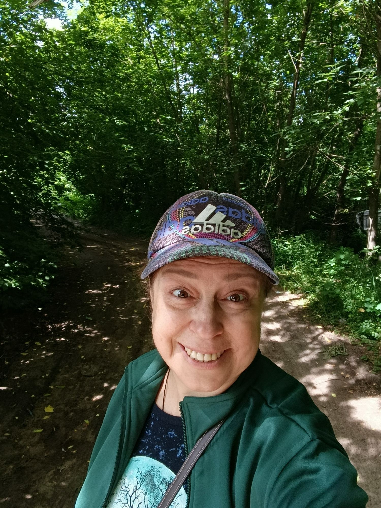 A selfie of a woman wearing a green jacket and a colorful Adidas baseball cap is taken outdoors on a dirt path surrounded by trees. The woman is smiling widely, with her eyes open wide and looking directly at the camera. The path appears to be in a wooded area, possibly a park or nature trail, with sunlight filtering through the leaves of the trees. The background shows dense foliage and a hint of a structure in the distance. The image was taken in the city of Vereya, Russia.