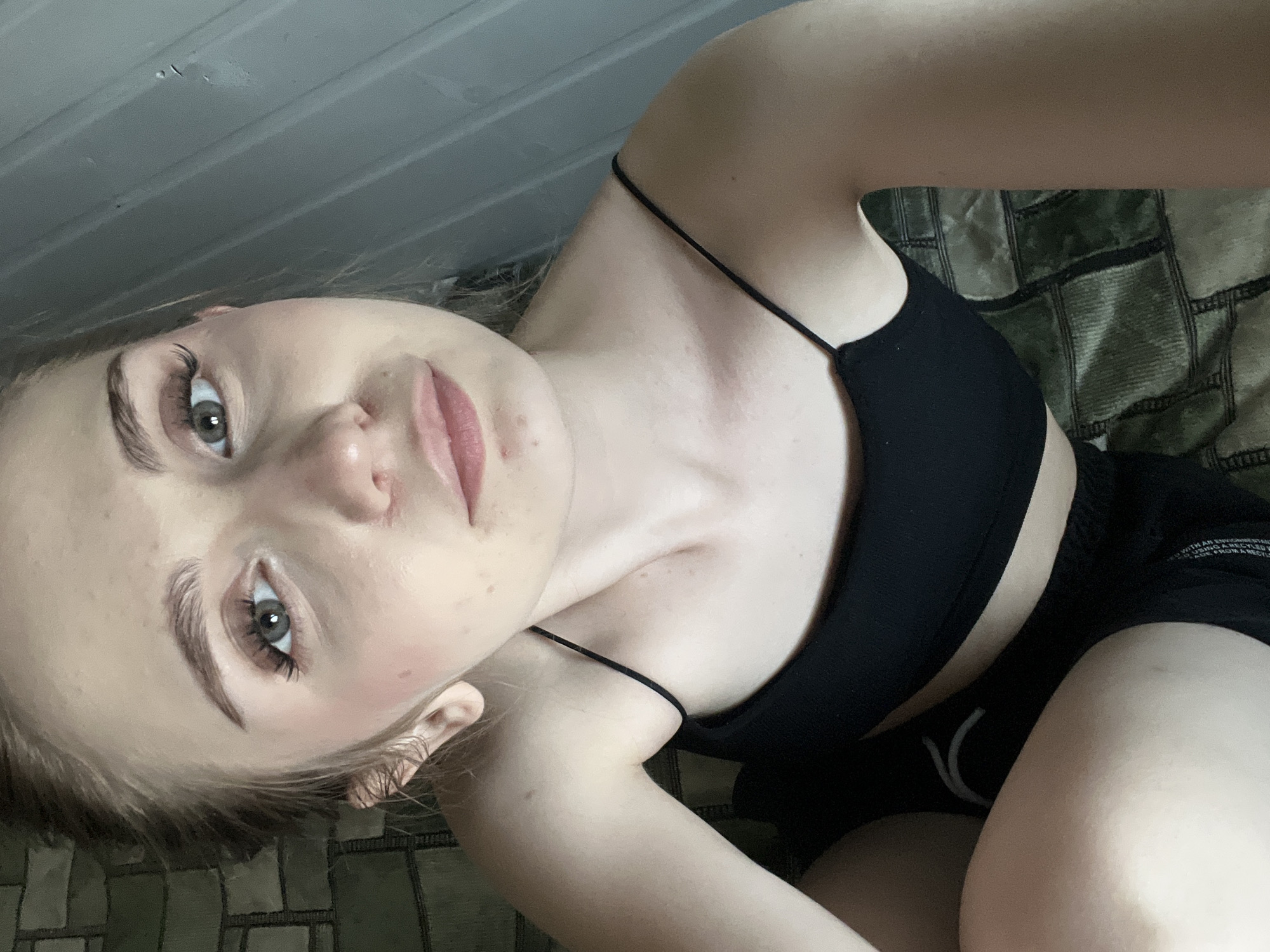 The media file is a selfie of a young woman with blonde hair and light skin, wearing a black sports bra and black shorts. She is lying down on a surface that appears to be a bed or couch covered with a patterned throw. Her head is tilted, and she is looking directly at the camera with a neutral expression. The background is dimly lit, with vertical wooden planks on the left side and a patterned fabric on the right. The woman has light-colored eyes, light eyebrows, and is wearing makeup, including mascara and lip gloss. There are some freckles visible on her face and chest. The overall mood of the image is casual and relaxed. There is no visible text in the media file. The provided location context is Ashkadar, Russia, but there are no specific visual cues in the image to confirm this location. The time of day and weather are not discernible from the image.