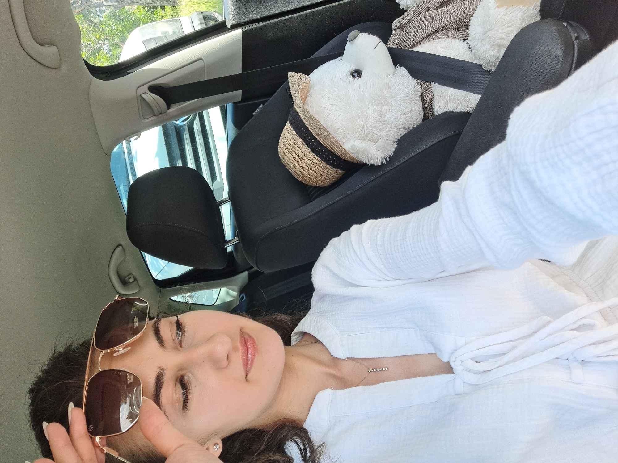 The image shows a young woman lying on the back seat of a car, holding sunglasses. She has dark curly hair and is wearing a white shirt and a necklace. A stuffed polar bear wearing a straw hat is also visible in the car, secured by a seatbelt. The setting appears to be a sunny day, with greenery visible through the car windows, suggesting an outdoor location, possibly Sofia, Bulgaria. The woman's expression is relaxed and content, indicating she is enjoying a car ride. The car interior, including the seats and seatbelt, are clearly visible, along with a rearview mirror and part of the car door. The overall atmosphere is casual and pleasant.