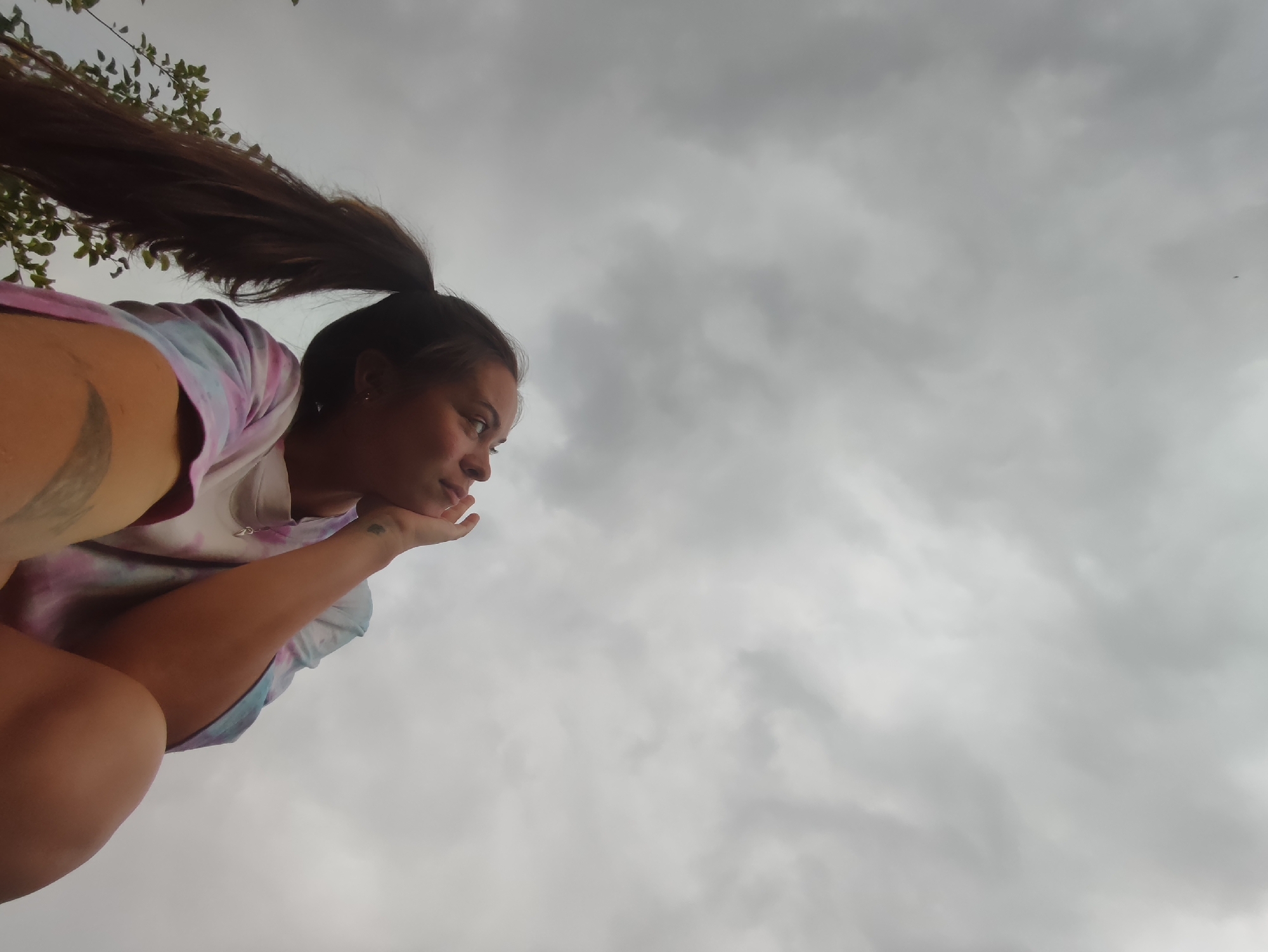 An extreme low-angle, wide shot captures a young woman with her long dark hair pulled back in a ponytail, which is being blown horizontally to the left by the wind. She is wearing a tie-dye t-shirt with pink, purple, and blue colors. Her left arm is bent and her hand rests on her chin as she looks to her right, out of the frame. A tattoo is visible on her upper left arm. The background consists of a cloudy, grey sky. No specific location within the city of Фоки, Russia is discernible from the image.