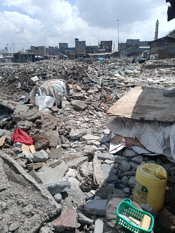 The image displays a vast landscape of demolition rubble and informal settlements in Nairobi, Kenya, specifically within areas like Mukuru kwa Reuben, following extensive demolitions along the Ngong River riparian land. The foreground and midground are covered by debris, including numerous grey concrete blocks, broken masonry, soil mounds, and assorted refuse. Prominent in the foreground are a piece of red fabric on the left, and a yellow plastic jerrycan alongside a green plastic basket containing a rectangular light-colored object on the right. A large, light-colored board, possibly wood or cardboard, stands upright, casting a distinct shadow. Mounds of debris are partially covered by white tarpaulins. In the background, a dense informal settlement composed of numerous single-story and multi-story structures made from corrugated metal, wood, and other scavenged materials extends across the horizon. A tall, slender mosque minaret is visible on the far right, rising above the settlement. A single streetlamp pole stands in the mid-distance to the right, and utility poles with wires are seen on the far left. The scene is brightly illuminated by natural sunlight under a partly cloudy sky, casting defined shadows. No people are visible.