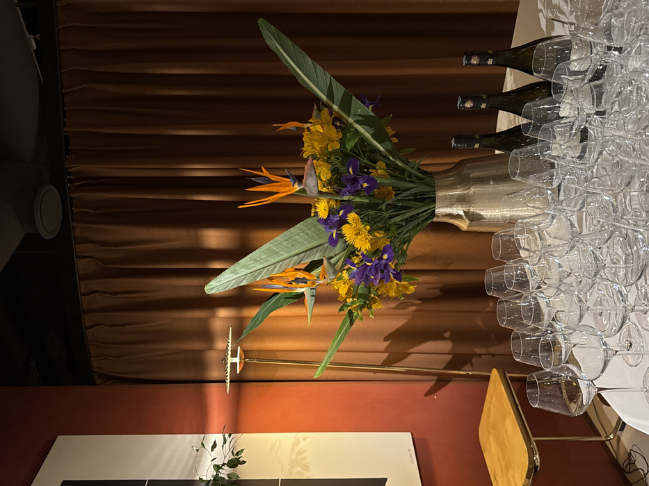 This image captures an indoor event setup within a venue in Yerevan, Armenia. A white-draped table in the foreground holds a dense array of clear, empty stemmed wine glasses, meticulously arranged for an upcoming gathering. To the right side of the table, three dark-bottled wines with light-colored labels stand upright. Centered on the table is a substantial floral arrangement housed in a textured, metallic gold vase. The arrangement consists prominently of orange Bird of Paradise flowers, deep purple irises, and a variety of yellow blossoms, interspersed with large green foliage. The background features a warm, reddish-brown painted wall on the left, partially displaying a large, minimalist black and white framed artwork. A tall, slender brass floor lamp with a disc-shaped diffuser casts a subtle glow near this wall. Below the artwork, a small, two-tiered table with a light-colored, possibly wooden, top is visible. The majority of the background is covered by heavy, pleated dark brown curtains. The ceiling is dark, revealing two circular ventilation grilles. No individuals, actions, or direct interactions are present in the scene.