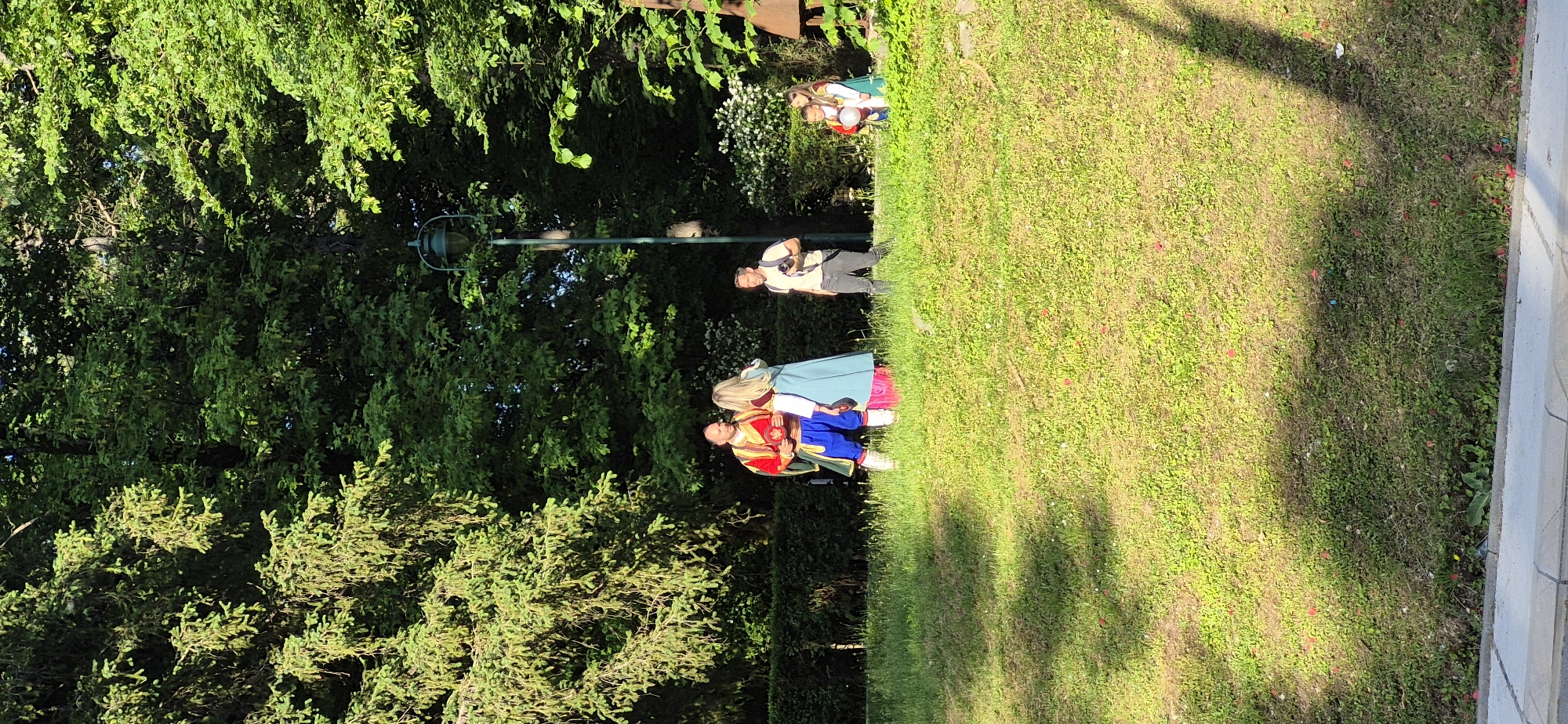 The image shows a scene in Cetinje, Montenegro. Two individuals in period costumes stand on a grassy lawn, adjacent to dense green foliage and a lamp post. One individual is dressed in a red, blue, and gold outfit, while the other is in a light blue and white ensemble with blond hair. A man with a camera is standing behind them. Further back, two other individuals in period clothing are also seen. A concrete curb and grass are present.