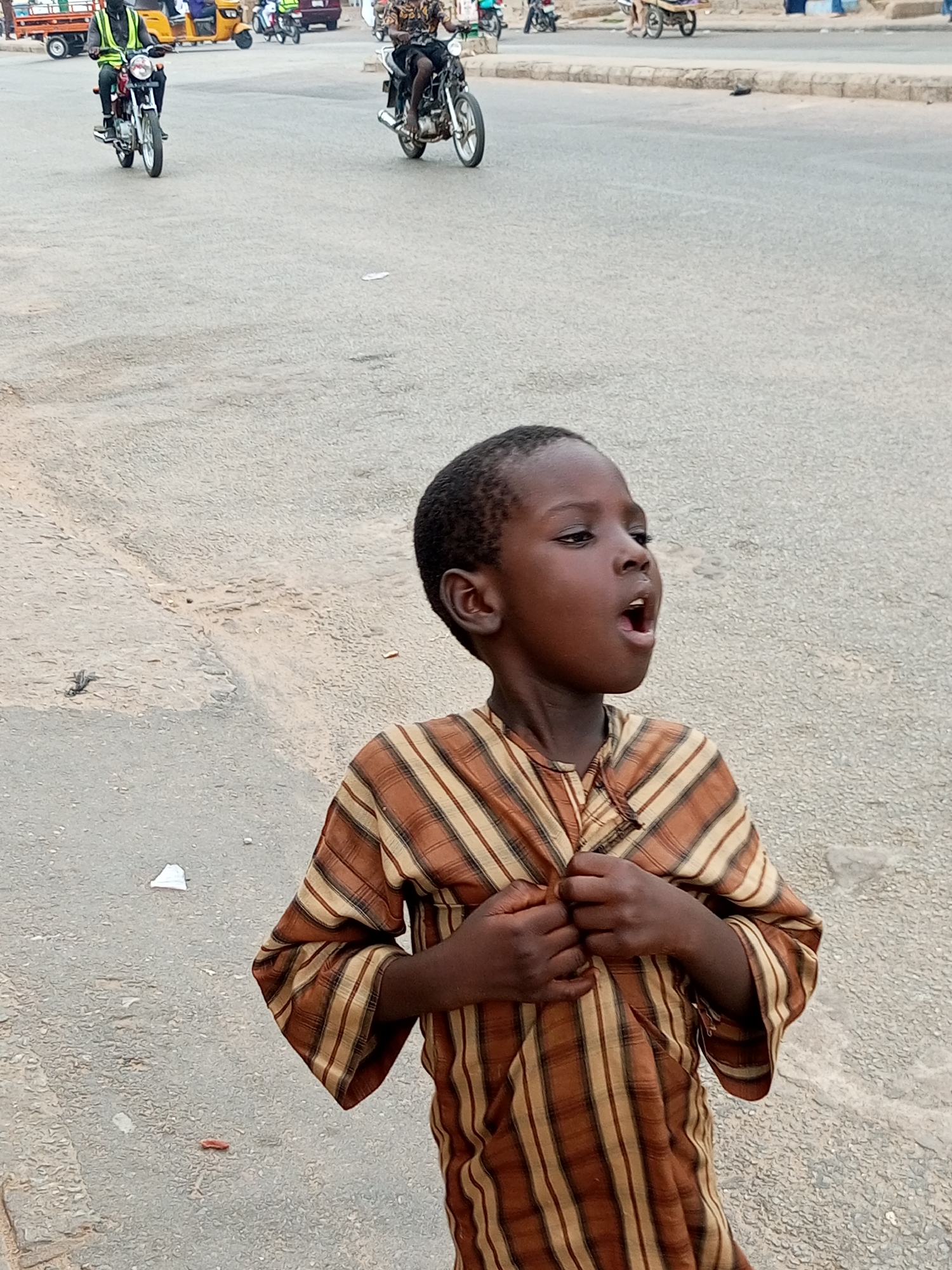In Arawa, Nigeria, a young boy with dark skin and short, dark hair stands on a paved road, looking up and to his right with his mouth open. He is wearing a brown and tan striped long-sleeved shirt. His hands are clasped over the front of his shirt. In the background, several motorcycles are visible on the road. One motorcycle in the left foreground has a rider wearing a bright green vest. Another motorcycle further in the background on the right has a rider wearing a dark shirt and black shorts. A three-wheeled vehicle is also present in the background to the left, along with a truck. The road surface appears to be asphalt.