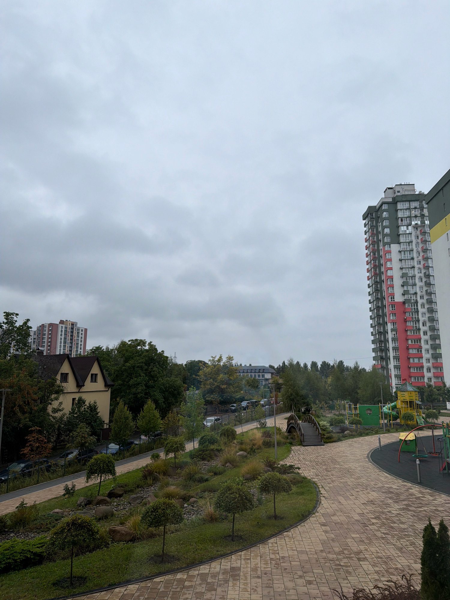 The image depicts an overcast day in a residential area of Kyiv, Ukraine. In the foreground, a paved walkway with a light brown, brick-like pattern curves through a landscaped area featuring meticulously pruned trees, rocks, and various grasses and shrubs. A small, wooden arched bridge spans a dry creek bed. To the left, a two-story residential building with a dark brown roof is visible, with parked cars along a road behind it. Further in the background, several multi-story apartment buildings rise above a line of trees. On the right side of the image, a tall, modern apartment building with a red, white, and green facade dominates the skyline. Adjacent to it, a playground area with colorful equipment, including slides and swings, is visible. The sky is uniformly gray and cloudy.