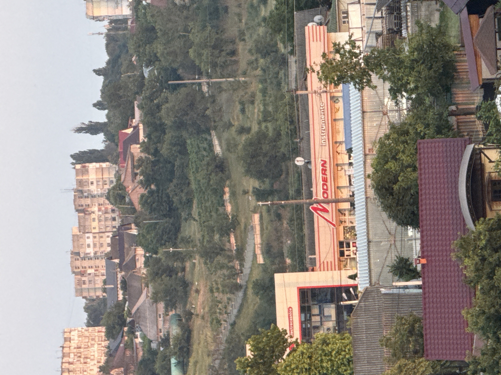 The image depicts a daytime, slightly hazy view of a residential and commercial area in Cahul, Moldova. In the foreground, a dark red tiled roof of a house is visible, with green trees and foliage surrounding it. Behind this, a corrugated metal structure and another house with a dark red roof are present.
In the mid-ground, a modern building with a red and white facade is prominent. It features large glass windows and signage that reads "electròcasnice" and "MODERN instrumente". Adjacent to this building is a structure with a metallic roof. Further back, more trees and greenery cover a sloping hill.
In the background, several multi-story apartment buildings with beige and pinkish facades dominate the skyline. These buildings appear to be of Soviet-era architecture. The overall environment suggests a mix of older and newer construction within a hilly terrain, characteristic of parts of Cahul. The sky is a pale, overcast blue. There are no people visible in the image.