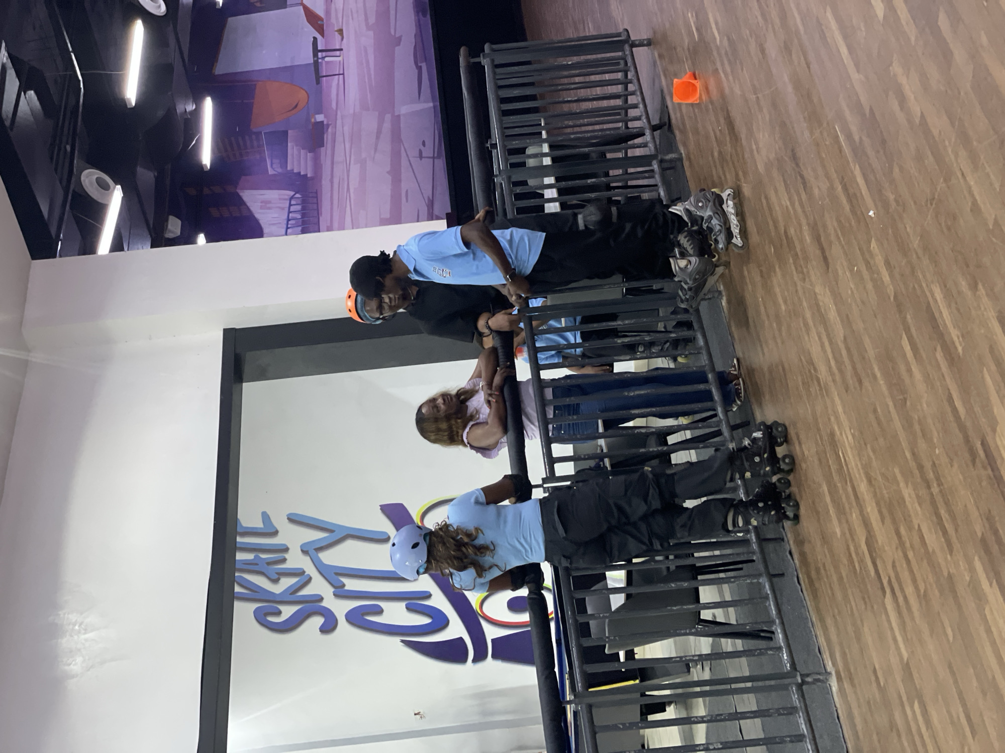The image captures an indoor roller skating rink, likely named "SKATE CITY" as indicated by the large blue and purple lettering on the white wall behind the subjects. The environment features a wooden floor, a black metal railing, and a large mural on the far wall depicting a purple cityscape with ramps and structures, possibly a skate park.
In the foreground, four individuals are visible. On the left, a woman wearing a light blue shirt, black pants, and a light blue helmet with roller skates is standing with her back to the camera, looking to her right. She is wearing black elbow and knee pads. To her right, a woman in a pink top and blue jeans is leaning on the railing, conversing with the people next to her.
A man in a black shirt and dark pants is standing with his back slightly turned, wearing an orange helmet and glasses. He appears to be speaking to another man to his right. This second man is wearing a light blue polo shirt with a logo, black pants, and roller skates. He is looking down and appears to be engaged in conversation.
In the background, another person in a blue shirt and black pants is seated on a chair. The scene is brightly lit by overhead fluorescent lights. A small orange cone is visible on the floor to the right of the railing. There are no immediate visual cues that definitively place this location within Moba, Nigeria, beyond the general context of an indoor entertainment venue.