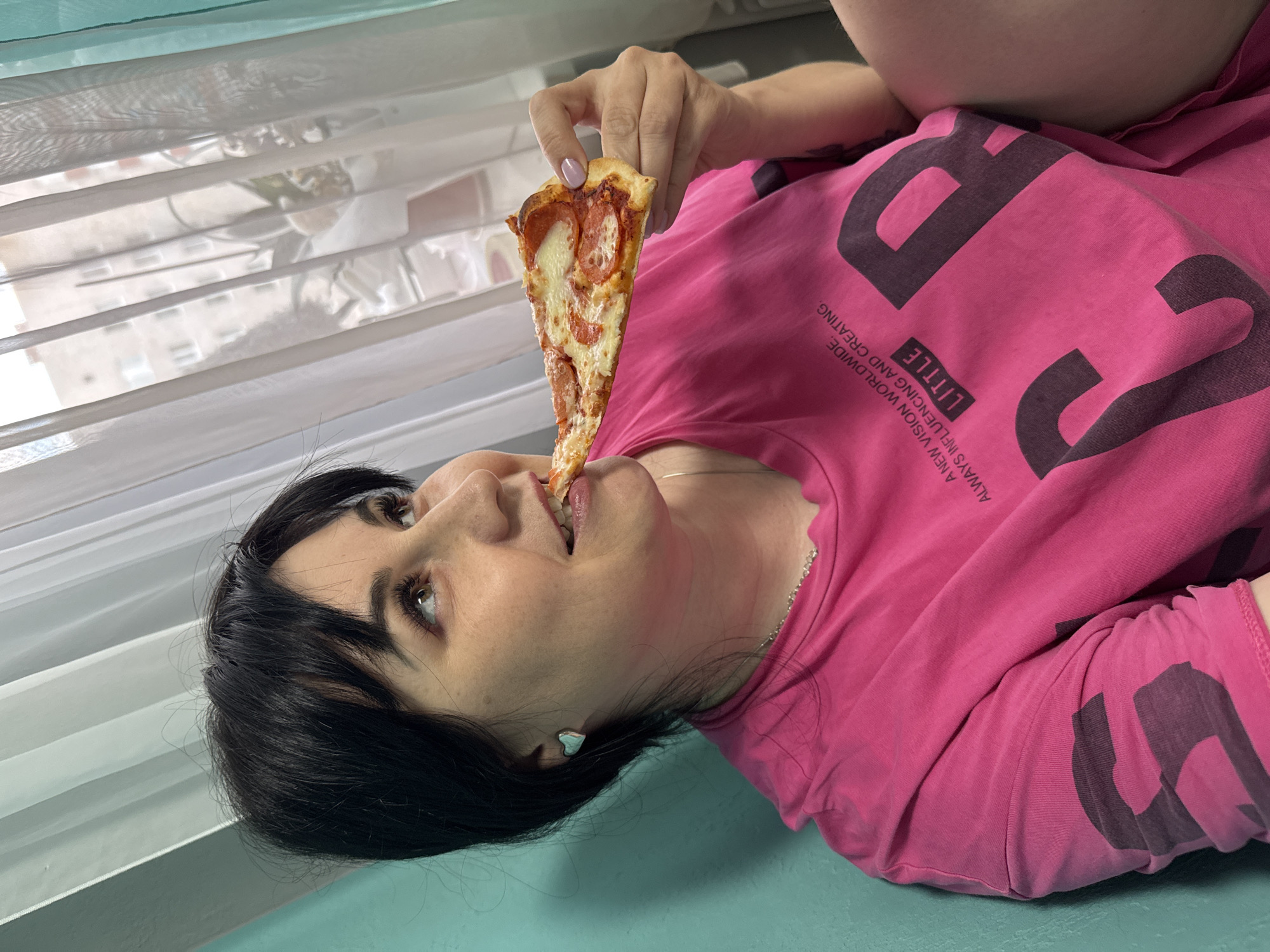 A woman with black hair is lying down, wearing a pink t-shirt, and eating a slice of pepperoni pizza. She is positioned next to a window covered by sheer white curtains. The location is Zav'yalovo, Russia.