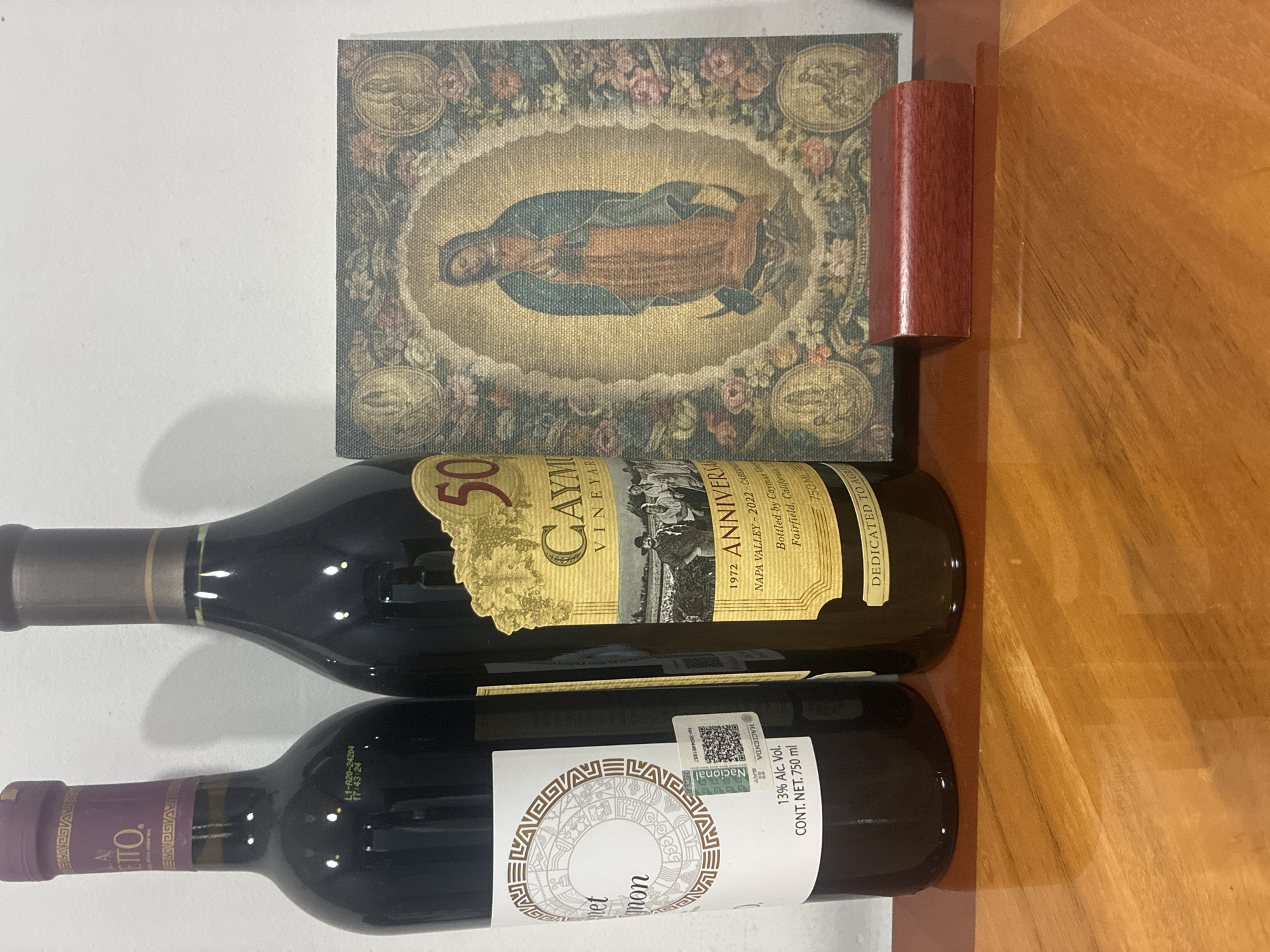 The scene consists of two wine bottles and a square artwork placed on a wooden surface. The top wine bottle has a golden label with "Caymus Vineyards 50 Anniversary" printed on it. The bottom wine bottle has a white label with an Aztec design and "Cabernet Sauvignon" written in the middle. Above the wine bottles, there's a square artwork depicting the Virgin of Guadalupe surrounded by flowers. A small red-brown wooden block sits next to the artwork. The background includes a white wall.
This specific location cannot be determined.