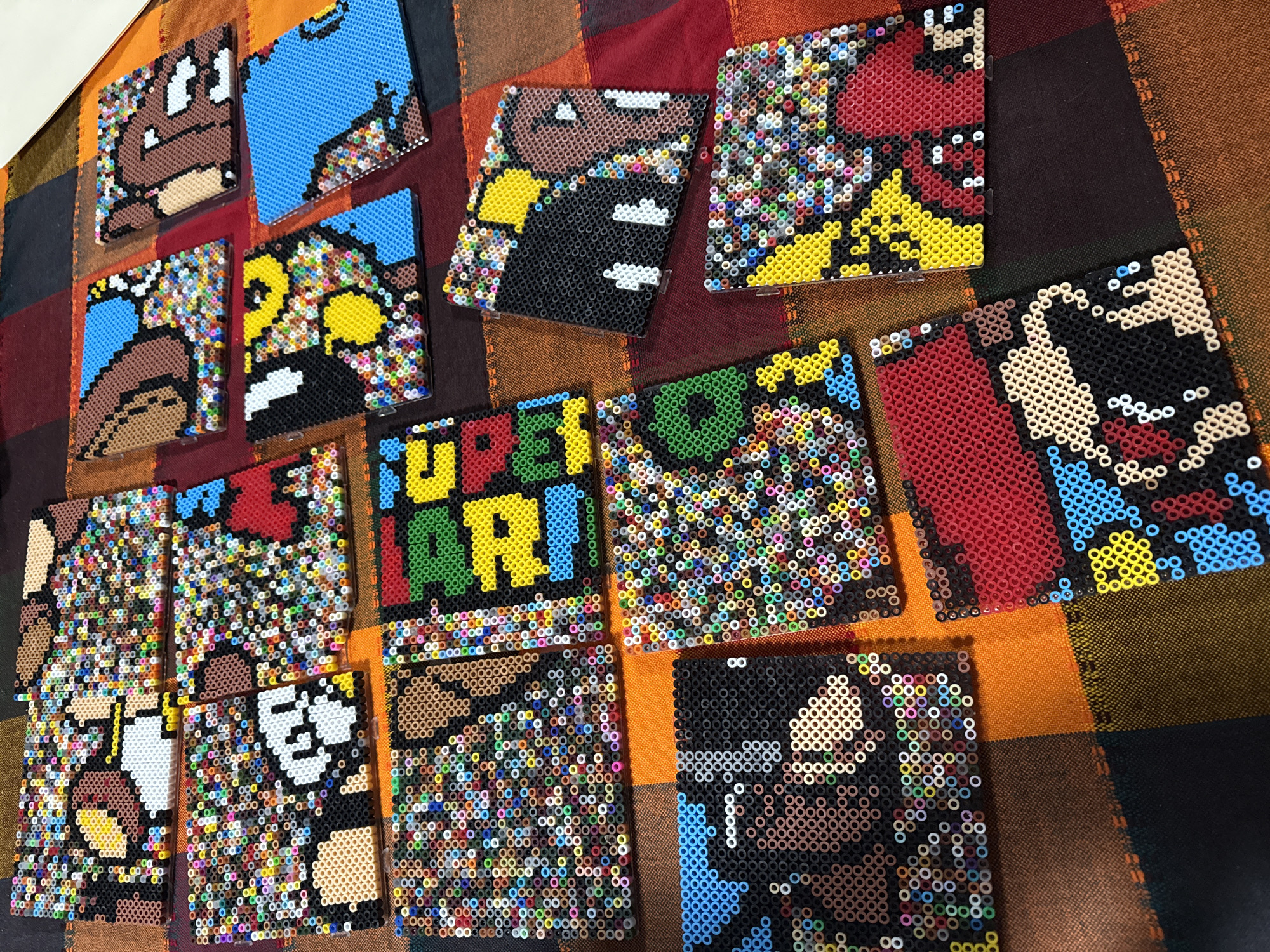 The image shows a collection of Perler bead art pieces laid out on a multicolored, possibly woven, textile surface. The designs feature characters and elements from the Super Mario video game franchise. Prominent characters include Mario, Luigi, a Goomba, and other iconic images like the "Super Mario" logo. The Perler bead artworks vary in size, mostly square or rectangular. The background textile has a pattern of varying colors, predominantly orange, brown, and black. The exact location in Puebla, Mexico, is not identifiable from the image content alone.