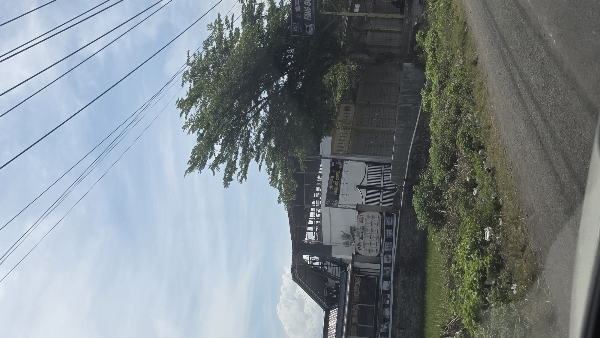 The image depicts a view of the Fans 86 Coffe establishment in Teungoh Baro, Indonesia. The building appears to be a multi-level structure with open sections and signage visible on its exterior. Adjacent to the building is a tree and a wall. A road runs alongside the structure, and utility lines are visible against a blue sky with clouds. Vegetation is also present along the road's edge. The photo seems to have been taken from a moving vehicle.