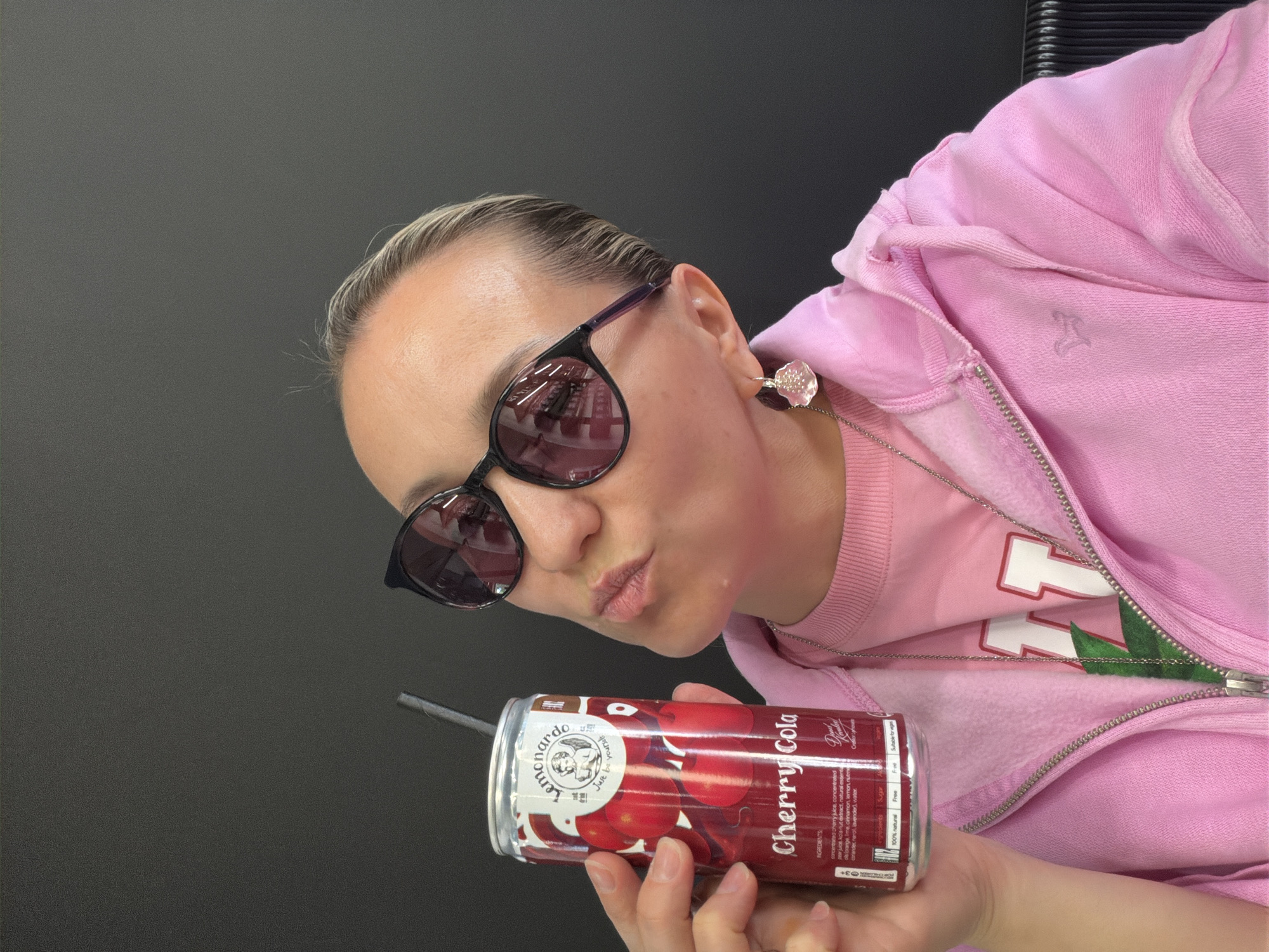 A woman wearing dark sunglasses and a light pink zip-up hoodie is holding a can of Leonardo Cherry Cola. The woman is facing the camera with a slight tilt of her head and a playful expression. The can of cola has a straw inserted. The background is a solid gray surface. The woman is wearing a silver necklace and a floral earring. The can's label indicates ingredients such as concentrated cherry juice, pear juice, and kola nut extract. There is no information about the location in the city of Izhevsk, Russia, in the image.