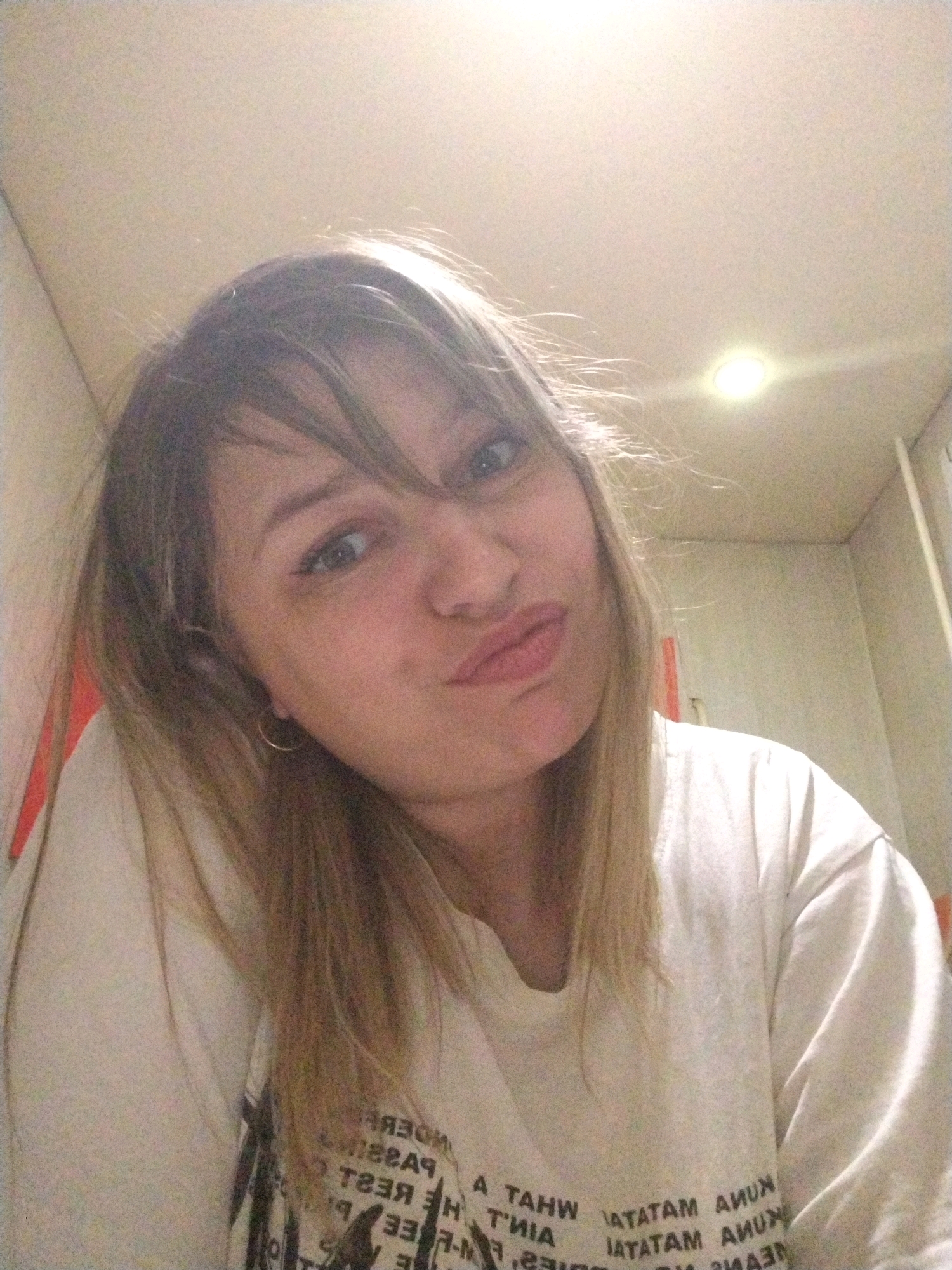 A young woman with blonde hair and bangs, wearing a light-colored t-shirt, is taking a selfie in an indoor setting. She is making a pouty face, with her lips pursed and her head tilted to the side. Her eyes are open and looking towards the camera. She is wearing a simple hoop earring on her visible ear. The background appears to be a wall with some indistinct markings and a light fixture on the ceiling. The lighting is somewhat harsh, creating a bright glow. The image is grainy, suggesting it was taken in low light conditions or with a lower quality camera. There is no identifiable context to place the image in Yaroslavl, Russia.