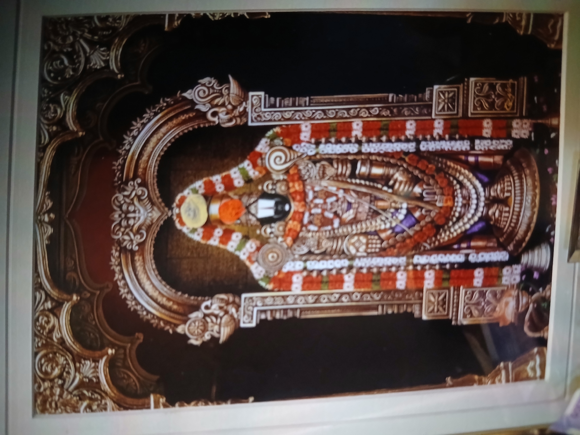 The image depicts a richly decorated statue of a Hindu deity, likely Lord Venkateswara (Balaji), positioned within an ornate, gilded frame. The statue is adorned with garlands of flowers in red, white, and green, along with various other colorful decorations. Offerings of what appear to be coins and colored powders are placed near the head. The background is dark, creating a stark contrast with the elaborate details of the statue and frame.
Based on the deity and ornamentation, the statue likely represents a deity from a temple in India. Specifically, this image appears to depict a representation of Lord Venkateswara, also known as Balaji. Lord Venkateswara is the presiding deity of the Venkateswara Temple located in Tirumala near Tirupati, Andhra Pradesh, India. The image is not specifically from Bengaluru.