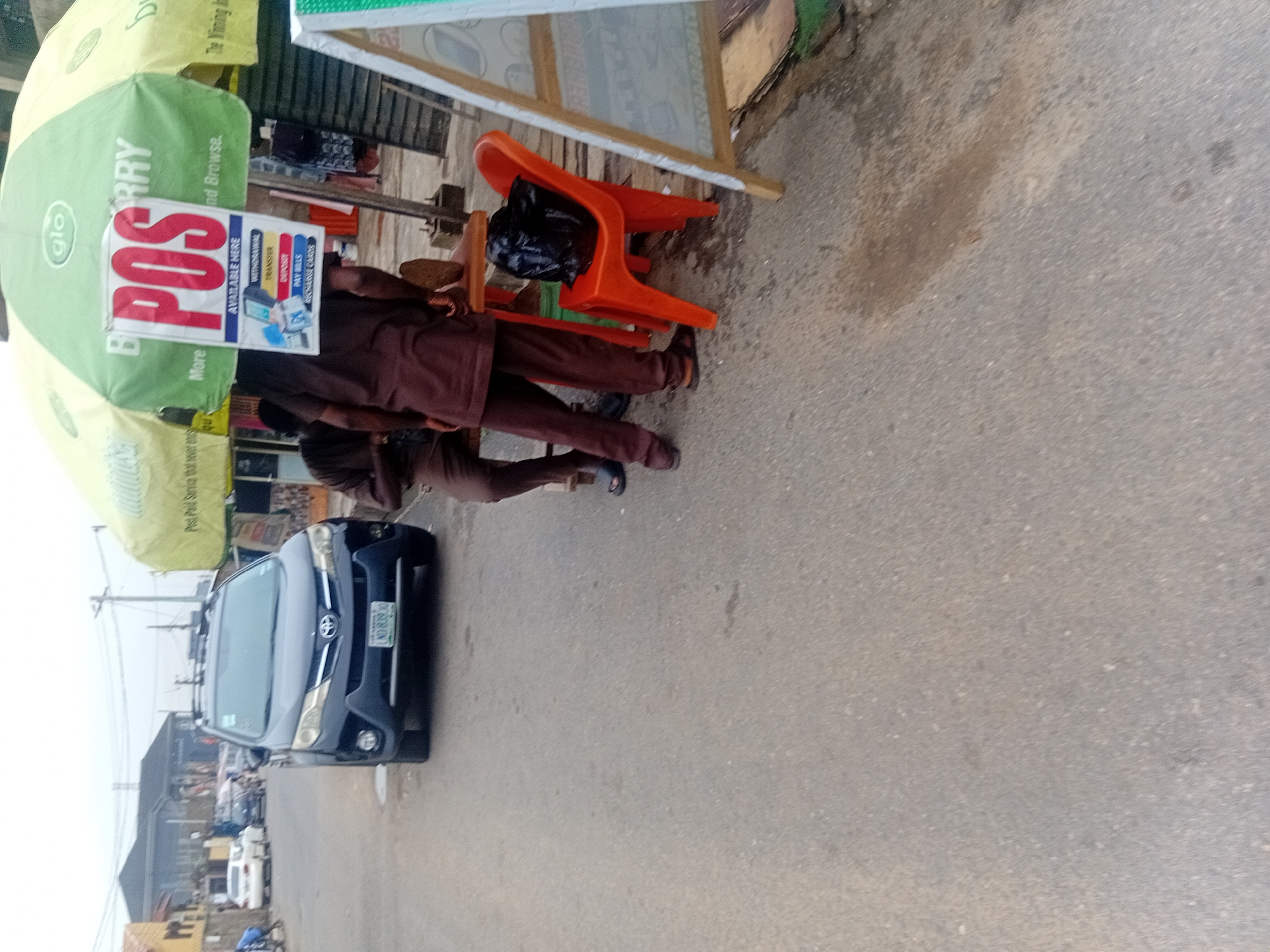 The image captures a street scene in Isheri, Nigeria, featuring an outdoor point-of-sale (POS) service. In the foreground, two individuals, dressed in similar brown outfits, stand near a green umbrella, under which the POS service is set up. One person appears to be interacting with the setup, while the other stands nearby.
A prominent white sign, suspended from the umbrella, boldly displays "POS" in large red letters. Below this, smaller text lists the services available: "AVAILABLE HERE", "WITHDRAWAL", "TRANSFER", "DEPOSIT", "PAY BILLS", and "AIRTIME CARDS". The green umbrella itself bears the "glo" logo, a Nigerian telecommunications company, and partial text like "Post Paid Service that never en..." and "nd Browse."
To the right of the people, an orange plastic chair is visible, with a black plastic bag placed on it. A dark blue Toyota car, with a Nigerian license plate "LND839 JD", is parked on the asphalt road to the left. The road surface shows some stains and wear. In the background, other cars and indistinct buildings suggest a busy commercial or residential area. The lighting indicates it is daytime, possibly under an overcast sky or during early morning/late afternoon, as there are no harsh shadows. The overall impression is that of a typical informal business setup common in Nigerian streets.