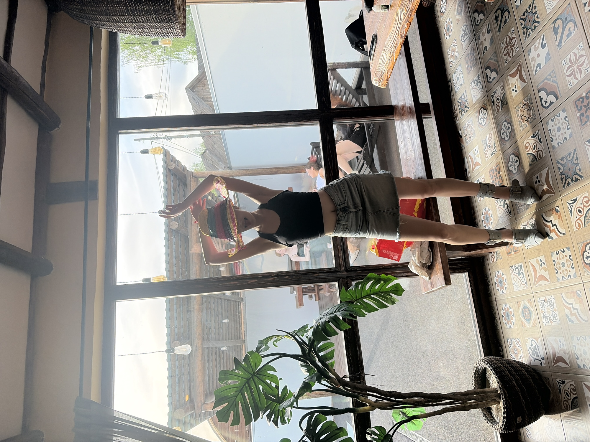 A woman wearing a black top, denim shorts, and grey sneakers is visible indoors. She is positioned near a large window with a brown frame, partially reflecting the outside scene of a location resembling Старое Михайловское, Russia. The window's reflection shows building structures, possibly shops, and vintage-style light fixtures. Inside, a plant with large green leaves stands near the window and decorative tiles line the floor.