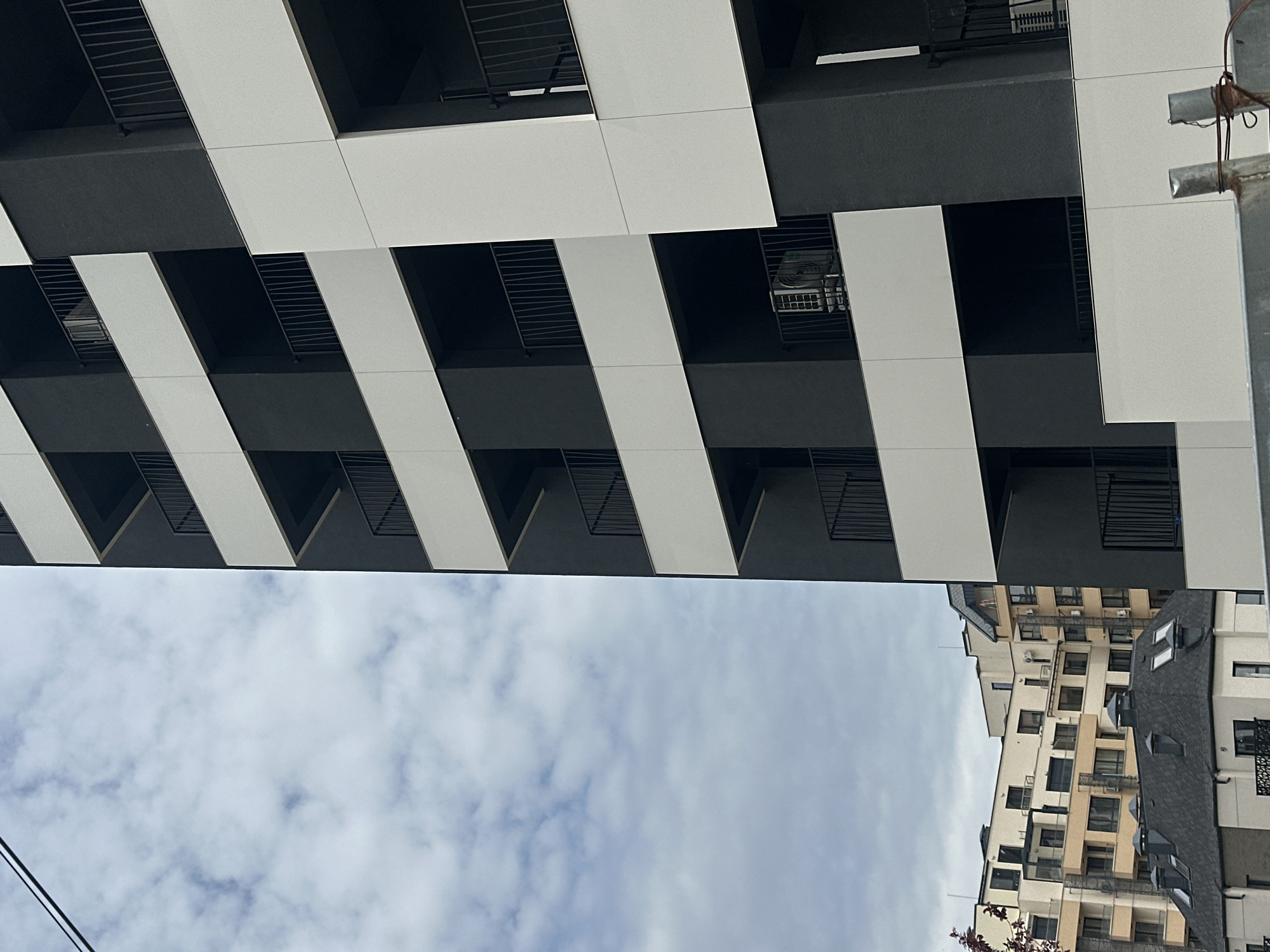 The image shows a view looking up at a building with a distinctive black and white striped facade. The building appears to have multiple balconies, indicated by the black recesses and metal railings. The sky is visible between the two buildings and is overcast with clouds. Another building is visible further away, with a more traditional facade and visible windows and balconies. The exact location in Sofia, Bulgaria is not discernible from the image. There are no people visible.