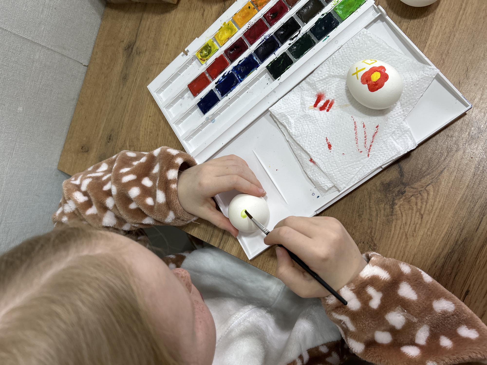 A child is shown from above, seated at a wooden table. They are wearing a brown and white polka-dot patterned fleece jacket or pajama top. The child is painting a white egg with watercolors. A palette of watercolors is situated next to the egg. One egg is already decorated with a red flower with a yellow center, a yellow plus sign, and a yellow halo shape. A paper towel with red paint streaks is below the decorated egg. The child is holding a paintbrush and painting a yellow circle on the plain white egg. Based on the environment, the photograph may have been taken in a residential setting. The city of Chaikovsky, Russia, is not visible in the image.