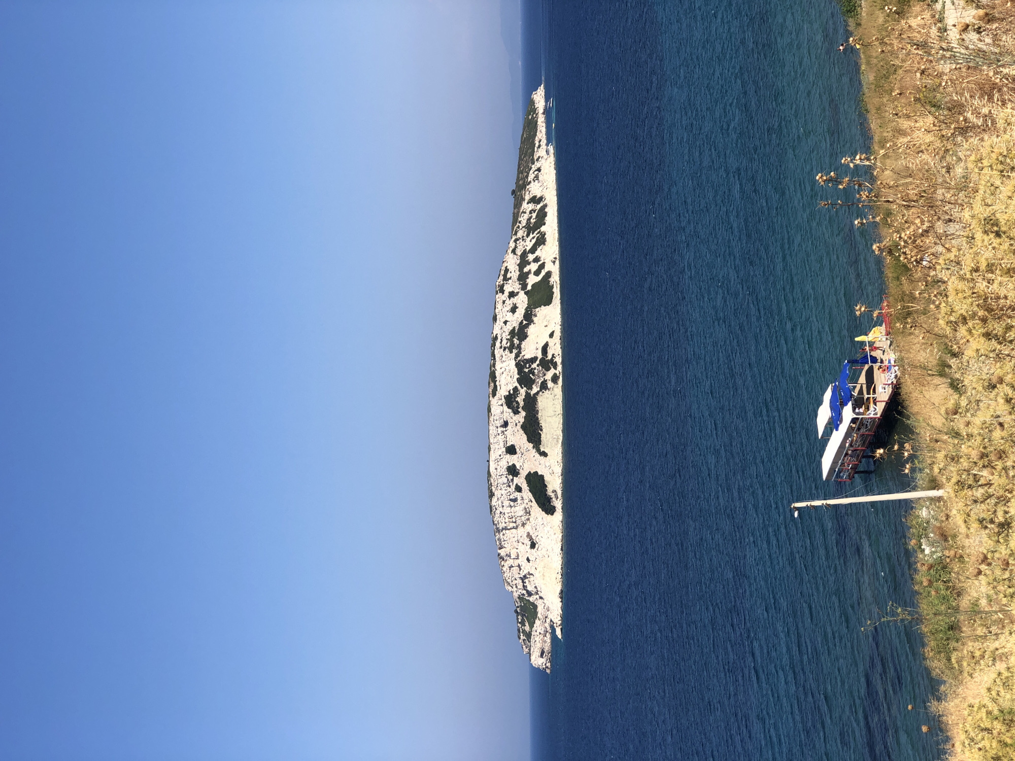 The image depicts a wide, clear blue sky above a calm, dark blue sea. In the distance, a large, rocky island with sparse green vegetation is visible. The island is illuminated by sunlight, highlighting its rugged terrain and white cliffs. Closer to the foreground, the sea meets a dry, grassy shoreline. A wooden pier extends from the shore, with a small covered structure on it and a boat moored nearby. A single lamppost stands on the shore next to the pier. The overall scene is serene and coastal. This location is in the vicinity of Karaburun, Turkey, a town known for its scenic coastline and islands.