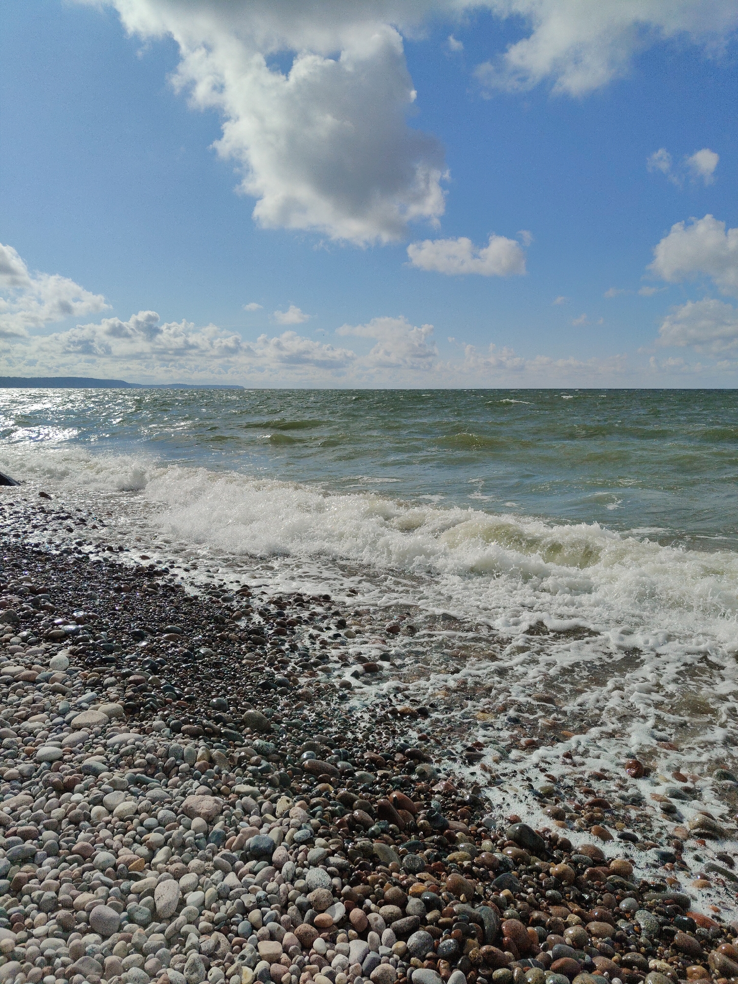 The image depicts a rocky beach on a sunny day. In the foreground, smooth, multicolored pebbles of various sizes cover the shore, with some wet from the incoming waves. White foam from the waves washes over the pebbles at the water's edge. The sea is a choppy, greenish-blue color, with waves breaking onto the shore. In the distance, a hazy shoreline with hills is visible. The sky above is a vibrant blue, dotted with fluffy white clouds. The scene appears to be a natural, unpopulated coastal area, consistent with a location like the beach in Pionersky, Russia, which is known for its pebble beaches and Baltic Sea coastline. There are no people or man-made structures prominently featured in the image.