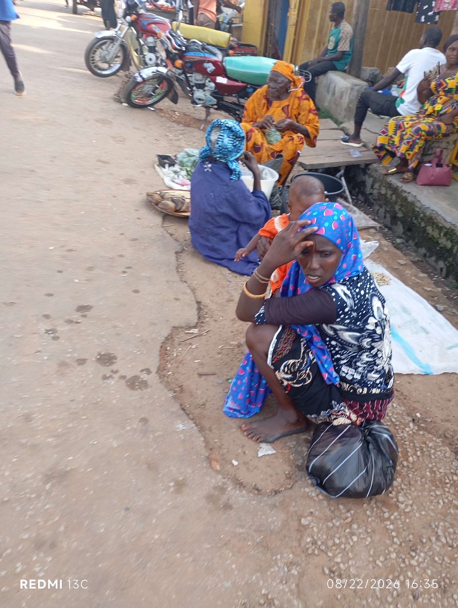 A street scene in Shendam, Nigeria, shows several people in an outdoor market setting. In the foreground, a woman wearing a blue headscarf and a patterned black and white dress is crouched down, holding her hand to her forehead. Next to her, a child in an orange shirt is visible. Further back, an older woman in a bright orange wrapper is sitting and appears to be knitting. Another woman in a colorful patterned dress is seated to her right, looking at something in her hand. Several motorcycles are parked in the background, and market stalls are implied by the presence of goods like fish and vegetables laid out on the ground. The ground is a mix of dirt and concrete. The time stamp "08/22/2026 16:35" and "REDMI 13C" are visible in the lower portion of the image.