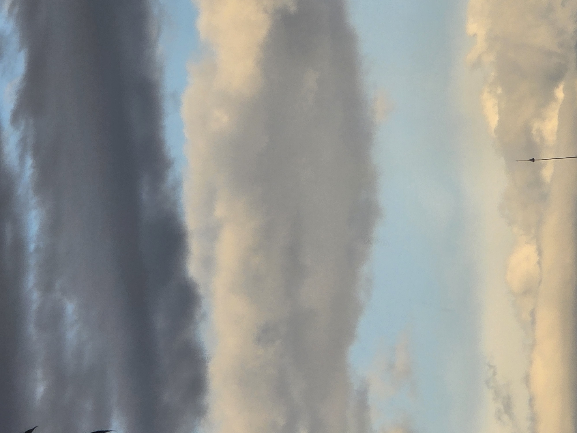 The image shows a vertical view of the sky in an urban environment, potentially in the city of Unknown, Unknown. Predominantly, the scene is filled with layers of clouds. On the left side of the frame, there is a dark, almost ominous band of grey clouds. Moving towards the right, the clouds lighten in color, transitioning into shades of grey and light yellow, giving the impression of sunlight filtering through. The sky peeks through in sections, appearing as a light blue. In the far right of the image, a thin, dark antenna-like structure is visible. The photograph's focus is largely on the sky and cloud formations, with the antenna being the only apparent man-made object.