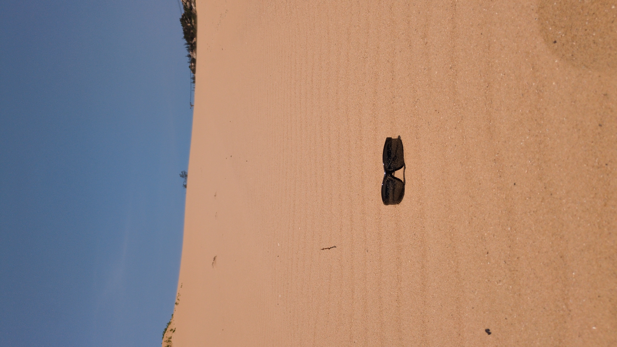 The scene is on a sandy beach in Phuoc Dinh, Vietnam. The sky is blue, and the sand is light brown. There is vegetation along the edge of the sand, on the left-hand side. A pair of dark-colored sunglasses lies on the sand.