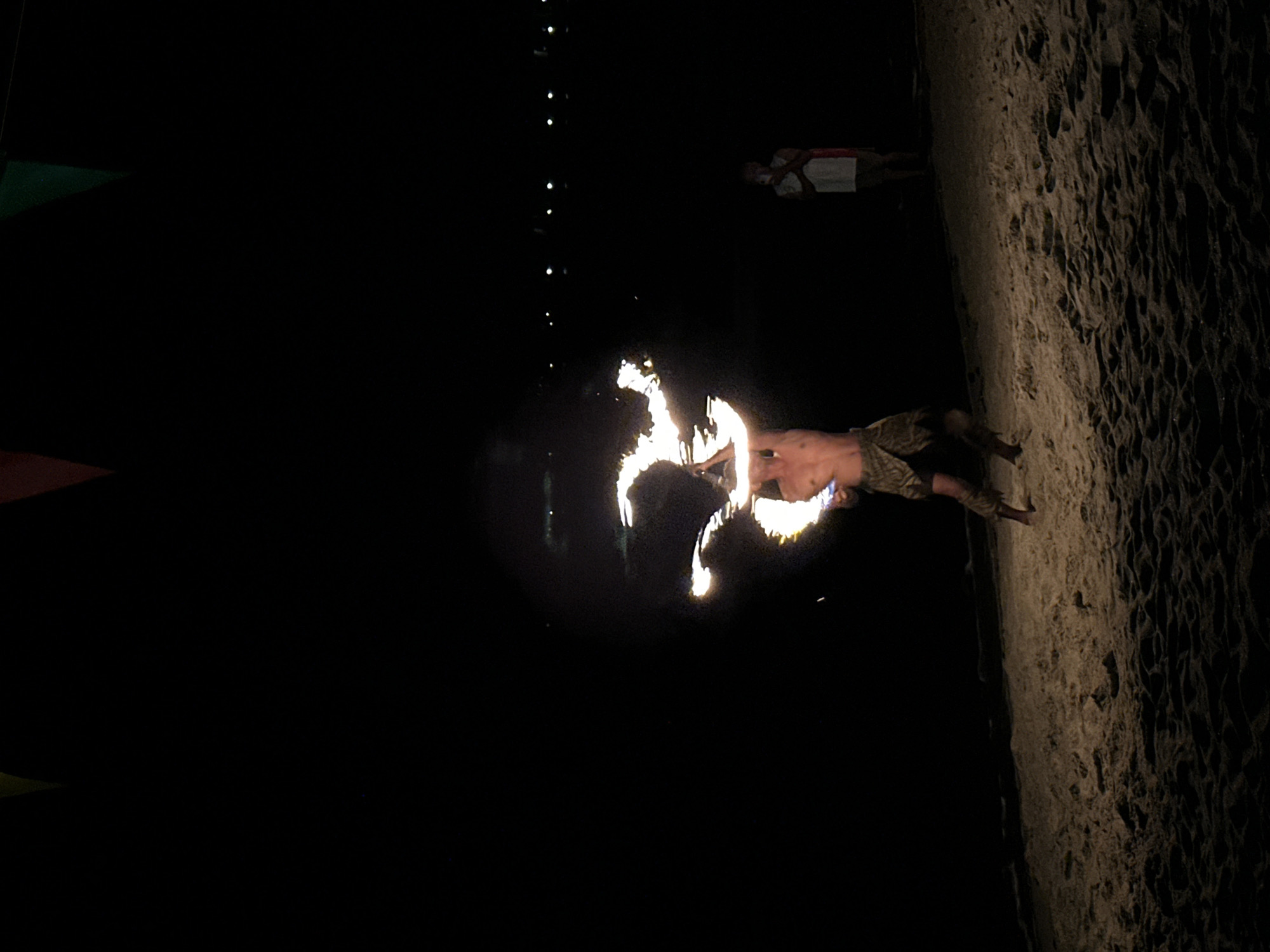 The image depicts a fire dancer performing on a beach at night. The location is Hat Khai Muk, Thailand. The fire dancer is shirtless, wearing camouflage shorts, and is wielding two flaming torches. A woman in a light-colored dress stands in the background. There are a few lights in the distance. The right side of the image shows the sand.