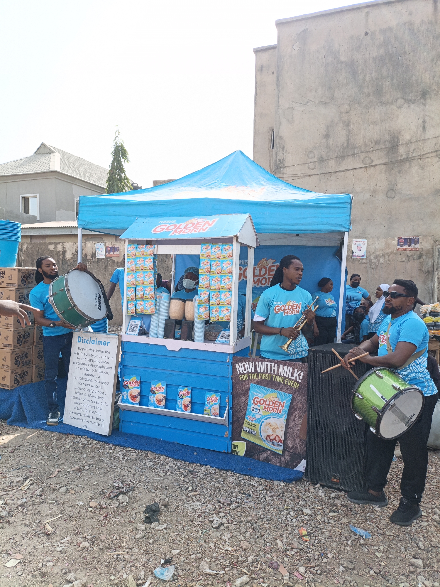 The image depicts a promotional event for Nestle's Golden Morn cereal, taking place in Kawaji, Nigeria. A blue tent and stand are prominently displayed. The stand features numerous packages of Golden Morn cereal, positioned on shelves and hung as decorations.
Several individuals are present, mostly wearing matching light blue t-shirts with the "Golden Morn" logo. Two men are playing musical instruments – one a large green drum and the other a brass instrument, likely a trumpet. One man stands next to the cereal stand wearing a mask. Other individuals are seated nearby, possibly staff or onlookers.
Stacks of boxes, presumably containing Golden Morn products, are visible to the left of the stand. A large speaker is next to the drummer. A sign on the stand includes a disclaimer. The ground is uneven and partially covered with litter. Background buildings are simple, concrete structures. Posters are hung on the adjacent wall behind the stand.