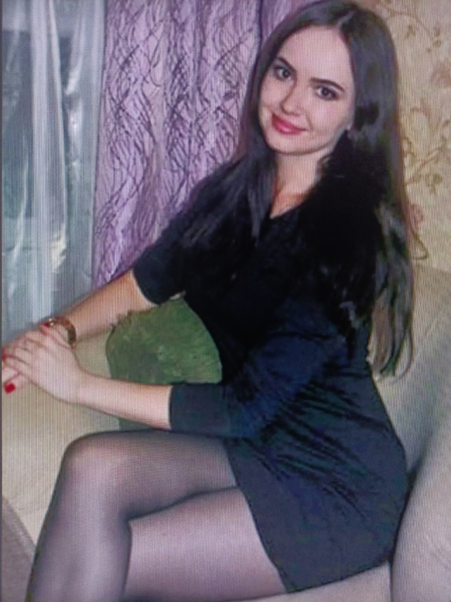 A young woman with long dark hair, fair skin, and dark eyes is sitting on a sofa, wearing a black dress and black tights. She is smiling, with red lipstick on her lips and red nail polish on her fingernails. Her right arm is resting on a green pillow. The background shows curtains and a patterned wallpaper. The image quality is low, with visible pixels and artifacts, suggesting it was taken with a low-resolution camera or has been compressed. The setting appears to be an indoor residential environment. There is no information in the image to suggest a location in Nyagan, Russia.