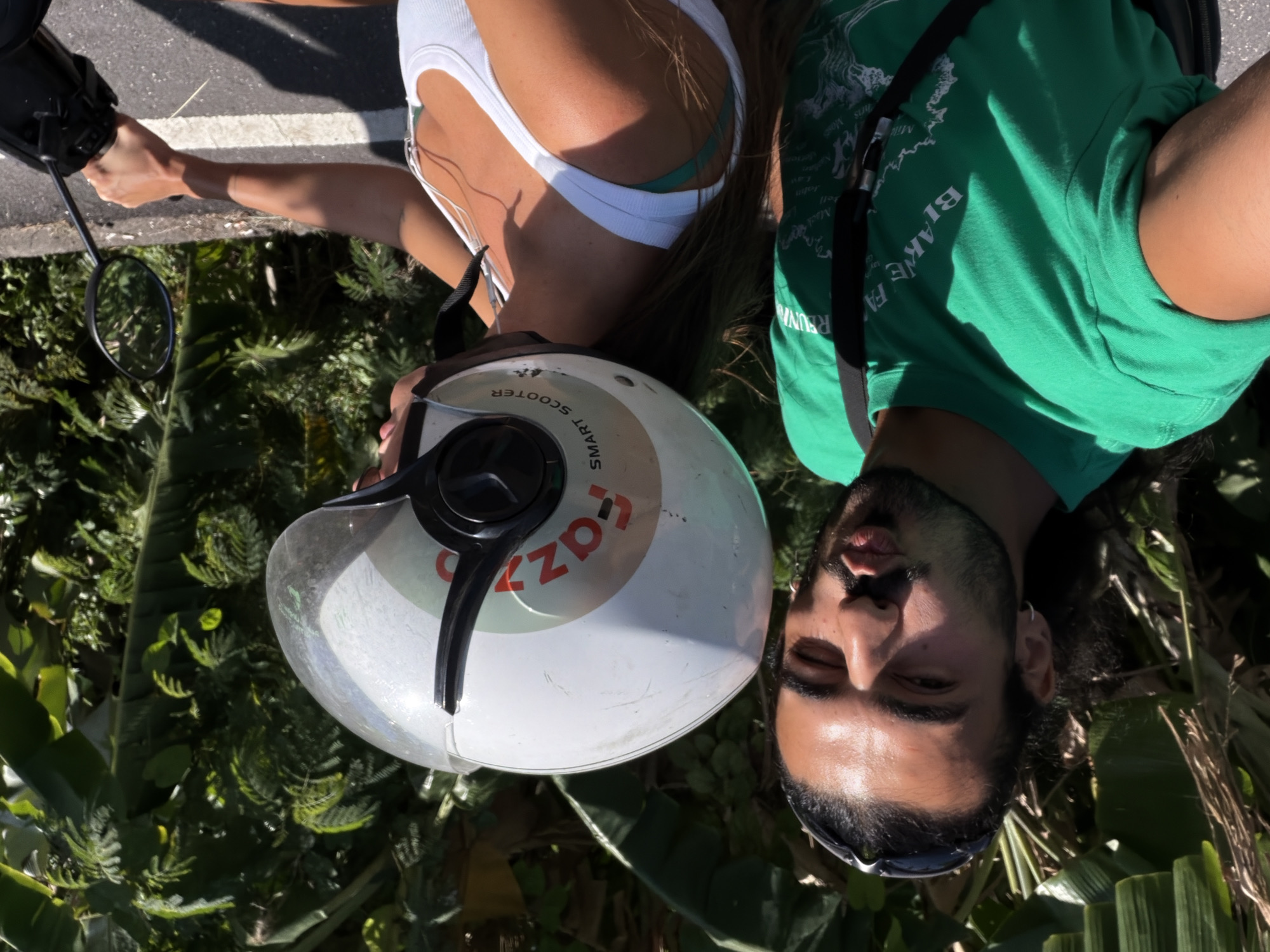 A man and a woman are riding on a scooter in North Kuta, Indonesia. The man is in the foreground, facing the camera and making a duck face. He is wearing a green t-shirt and sunglasses on his head. The woman is on the back of the scooter, wearing a white tank top and a helmet. The scooter is black and has a white sticker on the helmet that says "fazzo SMART SCOOTER". The background is lush and green, with palm trees and other tropical foliage. The road is paved and has a white line marking the edge. The lighting suggests it is daytime.