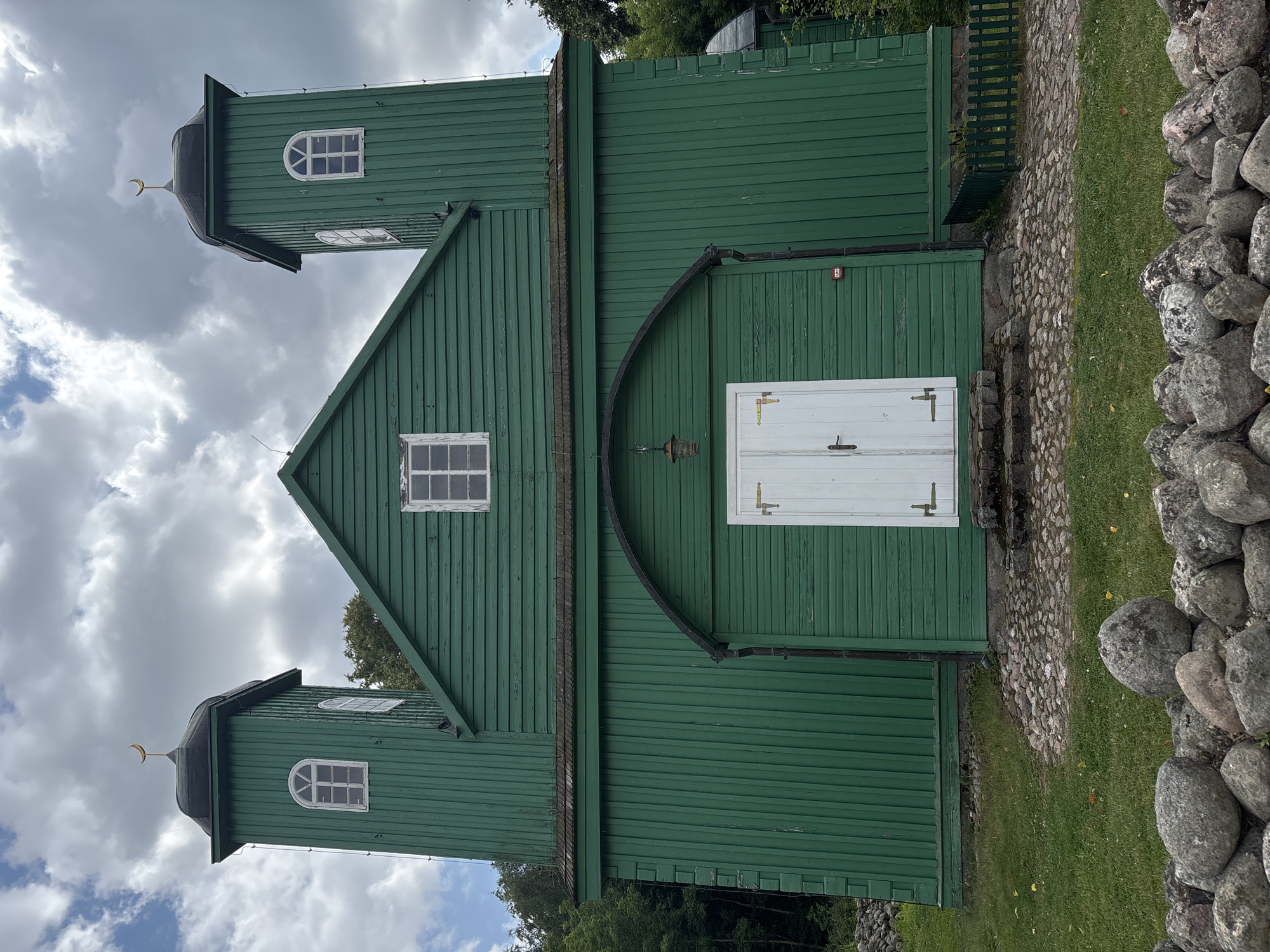 The image shows a bright green wooden mosque with two towers, located in Górka, Poland. The mosque has a central arched entrance with white double doors and a lantern hanging above. Each tower is topped with a black roof and a golden crescent moon. There are windows on the towers and on the main body of the building. The mosque is surrounded by grass, cobblestone paths, and a stone wall in the foreground. Trees are visible in the background. The sky is partly cloudy. There are no people visible in the image.