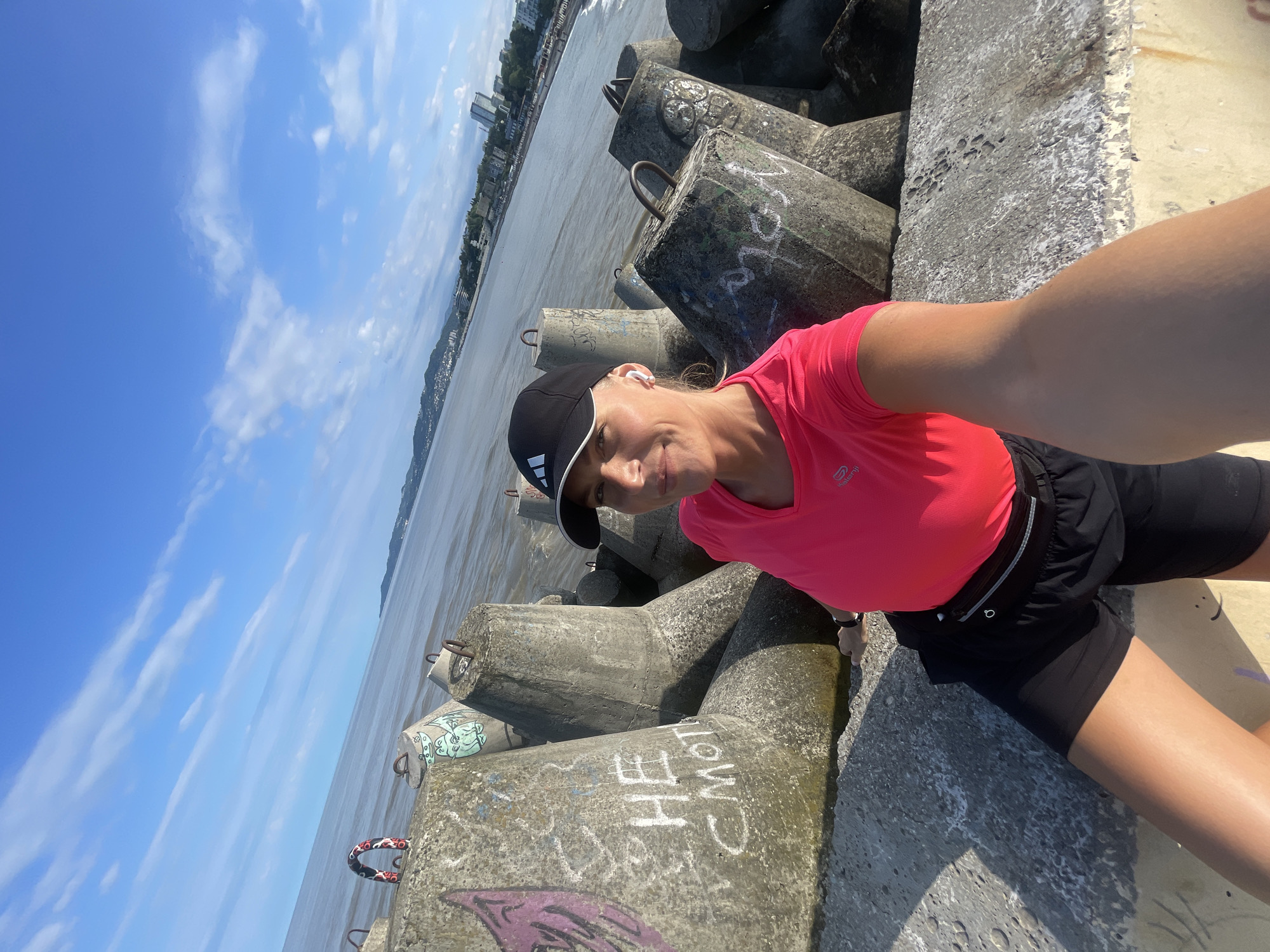 A woman in a pink t-shirt and black shorts poses for a selfie in Sochi, Russia. She's wearing a black Adidas baseball cap and seems to be resting on a concrete structure. The background shows the sea, the sky with clouds, and a distant city skyline. The concrete structure on which she's posing is covered with graffiti.