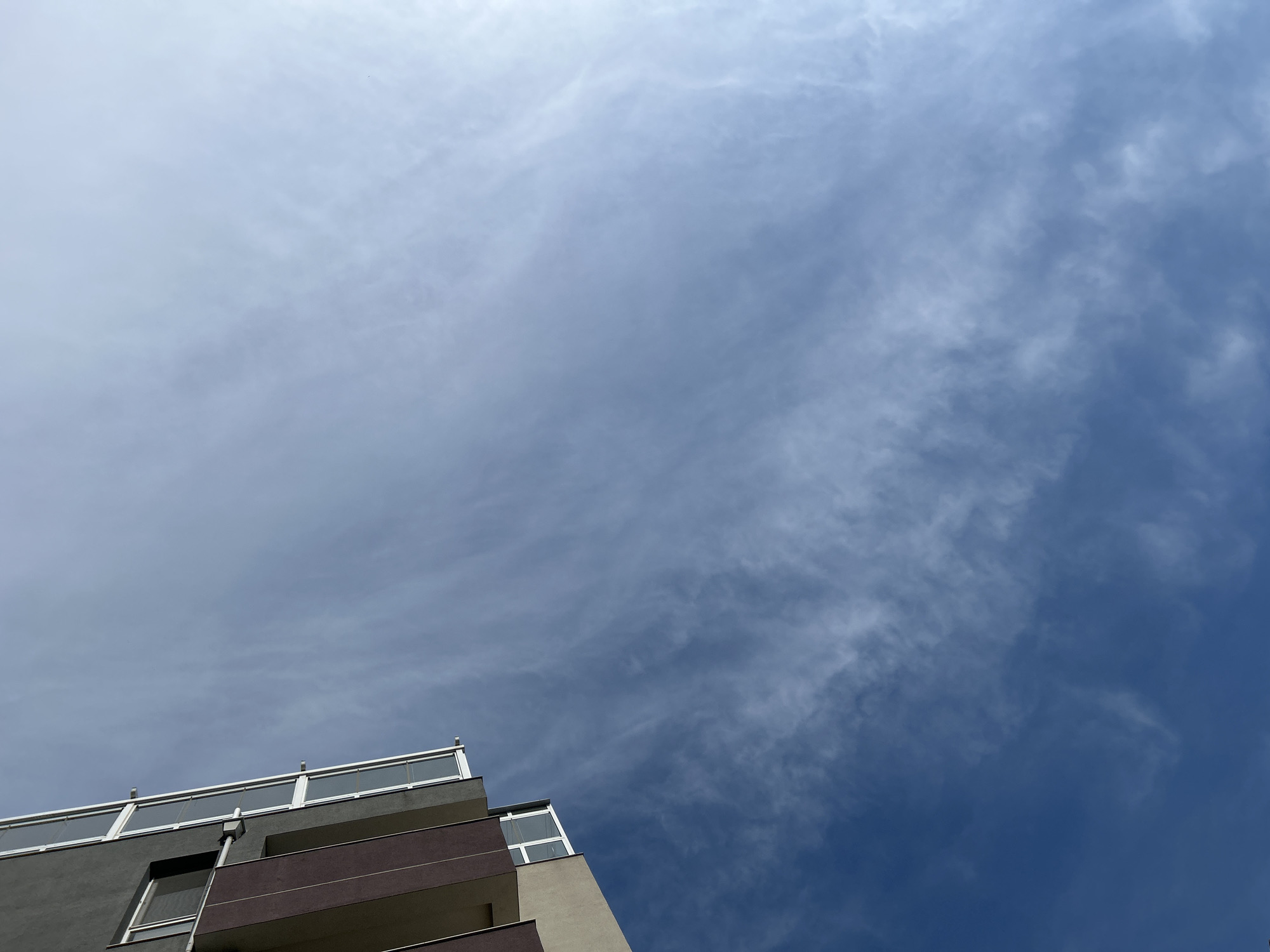 The image depicts a low-angle view of a modern building against a predominantly clear sky in the city of Varna, Bulgaria. The sky exhibits a gradient, transitioning from a deep blue at the bottom left to a lighter, almost washed-out blue-white in the upper right, consistent with direct sunlight originating from outside the frame. Scattered, thin, wispy cirrus clouds are visible across the expanse of the sky, presenting a streaked pattern, particularly noticeable in the central and upper portions. The building, a multi-story structure, occupies the upper-left quadrant, angled diagonally into the frame. Its upper visible section features a dark grey facade with a continuous white-framed glass railing, suggesting a balcony or a series of windows. Below this, a section with a reddish-brown, possibly terracotta, facade is present, followed by a lighter, neutral-toned section further down. A distinct white-framed window is visible on the lower part of the building's exposed wall. No individuals, animals, or specific actions are captured in the scene.