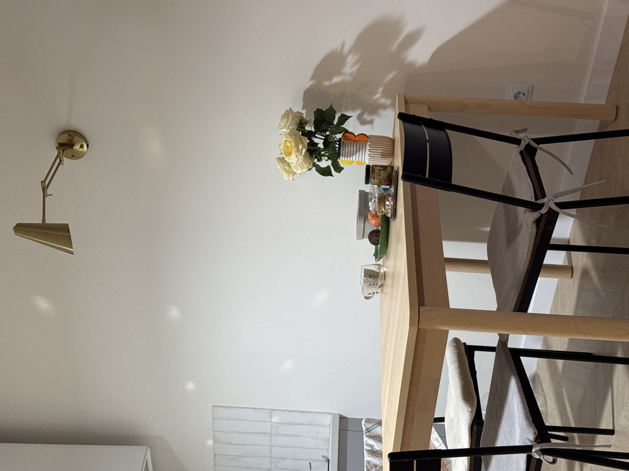 The image displays an indoor scene, likely a dining area or part of a kitchen, featuring a light wooden dining table and two chairs. The original image is rotated counter-clockwise.
**Subjects:**
The central subject is a light wooden dining table with a bouquet of white roses in a striped vase. Two chairs with black frames and light-colored fabric seat cushions, tied with white strings, are positioned around the table. Other items on the table include a clear glass mug with a pattern and small decorative or functional dishes. On the left wall, a brass-toned adjustable wall lamp is mounted. In the background on the left, a section of a kitchen counter with white subway tiles is visible. A white electrical outlet is partially seen on the right wall.
**Activity or event taking place:**
The scene depicts a static interior space; no explicit activity or event is occurring.
**Notable details:**
A prominent shadow of the roses and their foliage is cast on the white wall behind the table, suggesting a bright light source, likely artificial, from the lower left. The lighting appears to be indoor, possibly daytime or evening with artificial illumination. The setting is a contemporary domestic interior. No specific location cues are visible to confirm the provided context of Izhevsk, Russia.
**Visible text:**
There is no discernible text visible in the image.