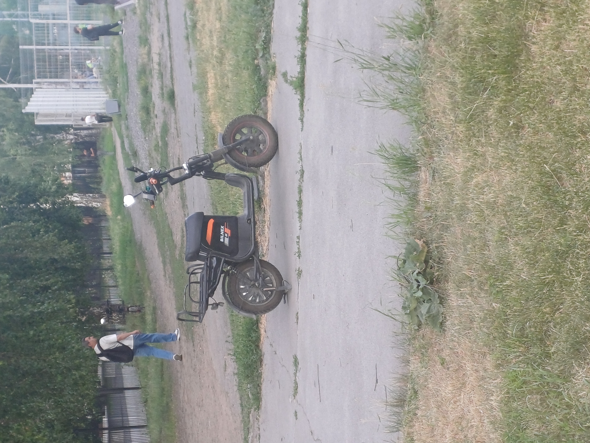 A man in a white t-shirt and blue jeans is standing next to a black electric scooter. He is wearing a dark backpack and has a casual demeanor. The scooter has a storage rack at the back and the brand name "BILNEX" is visible on its side. The scene appears to be outdoors, with a paved pathway and a grassy area. In the background, there are trees, a fence, and some structures that might be part of a park or a recreational area. There are also other people visible in the distance, some of whom seem to be near a fence or structure. The lighting suggests it is daytime, possibly late afternoon or early evening. The overall atmosphere is relaxed and casual, hinting at a leisurely activity or a moment of pause. The location context is Novosibirsk, Russia.