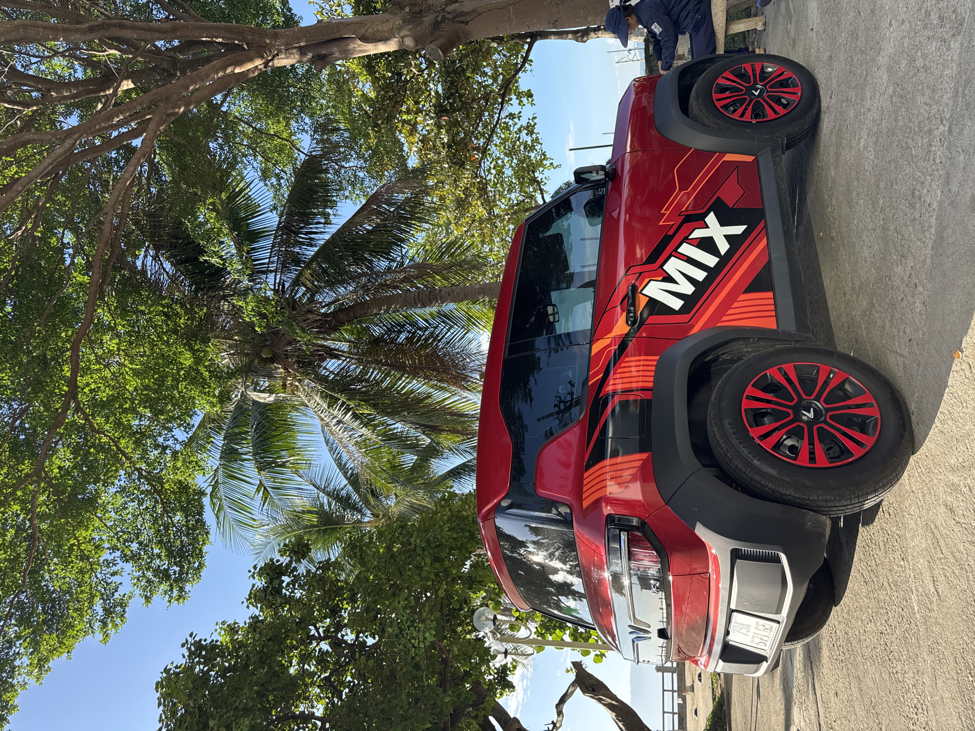 A red electric car with "MIX" emblazoned on its side is parked on a paved area beneath trees in Nha Trang, Vietnam. The car has a prominent graphics package featuring black and orange geometric patterns. A person wearing a blue cap and jacket sits on a bench to the right of the car, looking down. In the background, palm trees and other foliage suggest a tropical coastal environment, with the sky visible through the leaves. The sea and a distant coastline are visible beyond a low wall in the background. The license plate of the car reads "79A 571.82".