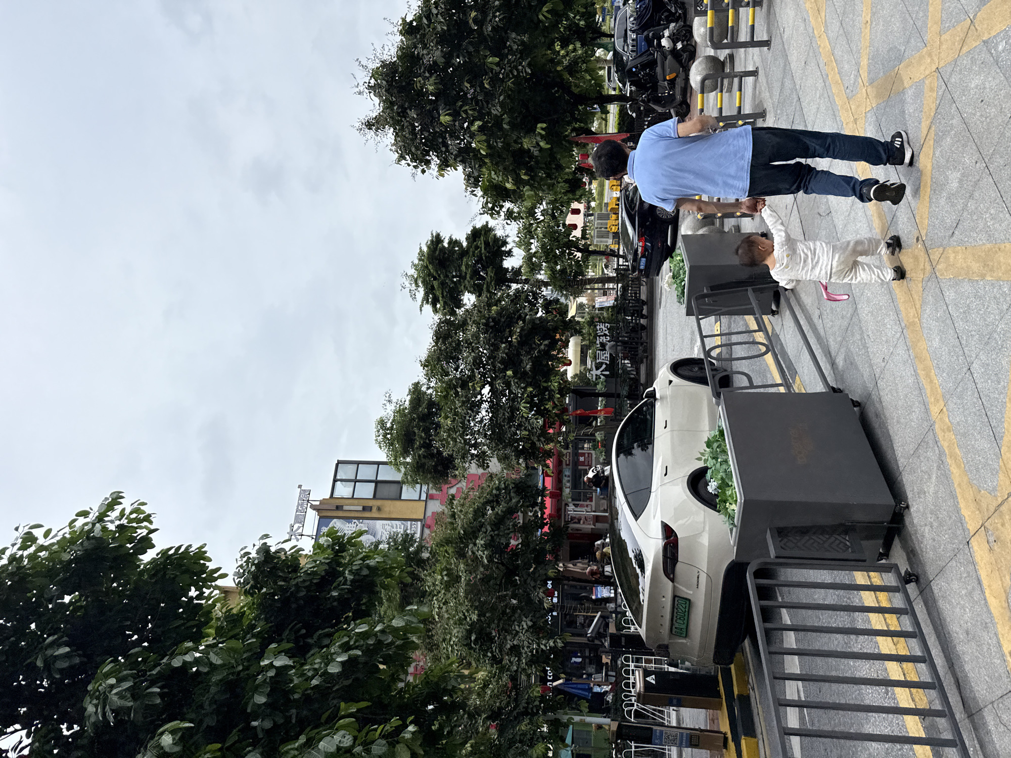 A man in a blue polo shirt and dark jeans walks hand-in-hand with a toddler in a white pajama set down a sidewalk in Haizhu District, China. The child holds a pink ribbon in their free hand. A white Tesla Model Y is parked on the street to the left of the sidewalk, with a green license plate reading "B A C60220". Buildings with signage are visible in the background, along with trees lining the street. The sky is overcast.