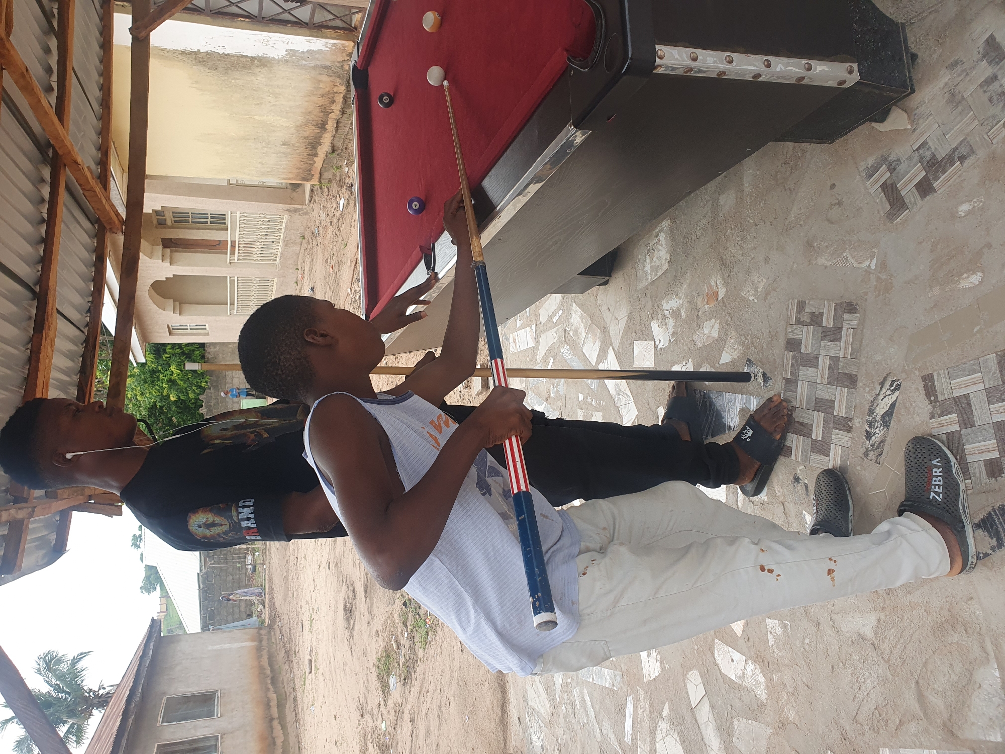 Two young Black males are playing pool outdoors in Lokoja, Nigeria, under a simple, open-sided shelter with a corrugated metal roof supported by wooden beams. The shelter's floor is a mix of sand and scattered square tiles. In the background, a modest, light-colored residential building with arched doorways and windows is visible.
The male on the right, wearing a black t-shirt with a graphic design and white earbuds, stands behind the pool table. The male on the left, in a white tank top and light-colored pants, is actively playing. He is leaning over the red felt pool table, holding a cue stick with a blue and red striped handle. The pool table has several balls on it, including a purple ball, a white cue ball, an orange-numbered ball, and a black 8-ball. The ground around the table is dusty, with some scattered debris. The overall environment suggests a casual, possibly informal recreational area.