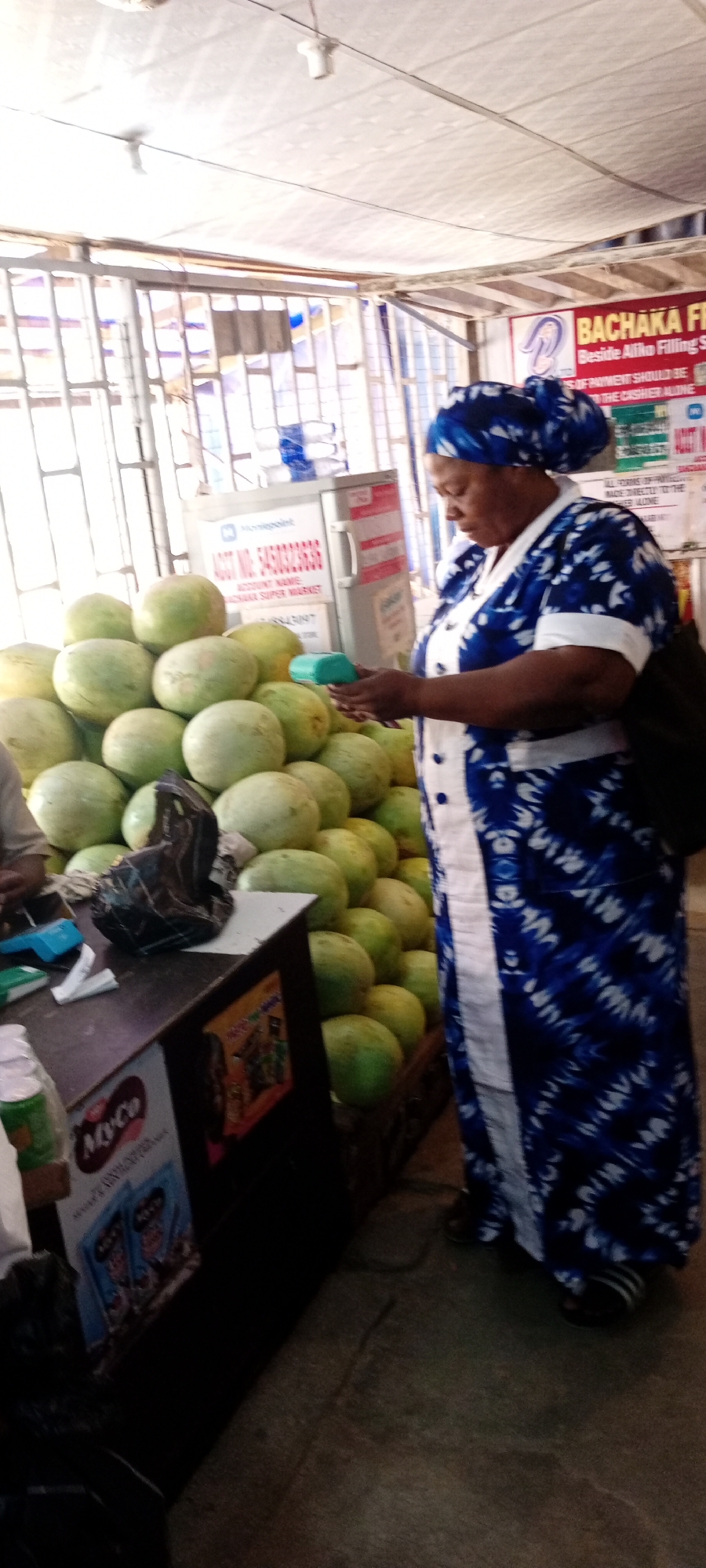 An image shows a woman in a blue and white patterned dress and headscarf holding a small green electronic device, possibly a payment terminal, while standing in front of a large pile of green watermelons. To the left of the watermelons, a dark counter displays some products, including a "Myco" branded item. In the background, a white refrigerator-like appliance with signage, a metal grate structure, and what appears to be a white tarp ceiling are visible. The scene suggests a marketplace or roadside stall in Aviation Quarters, Nigeria.