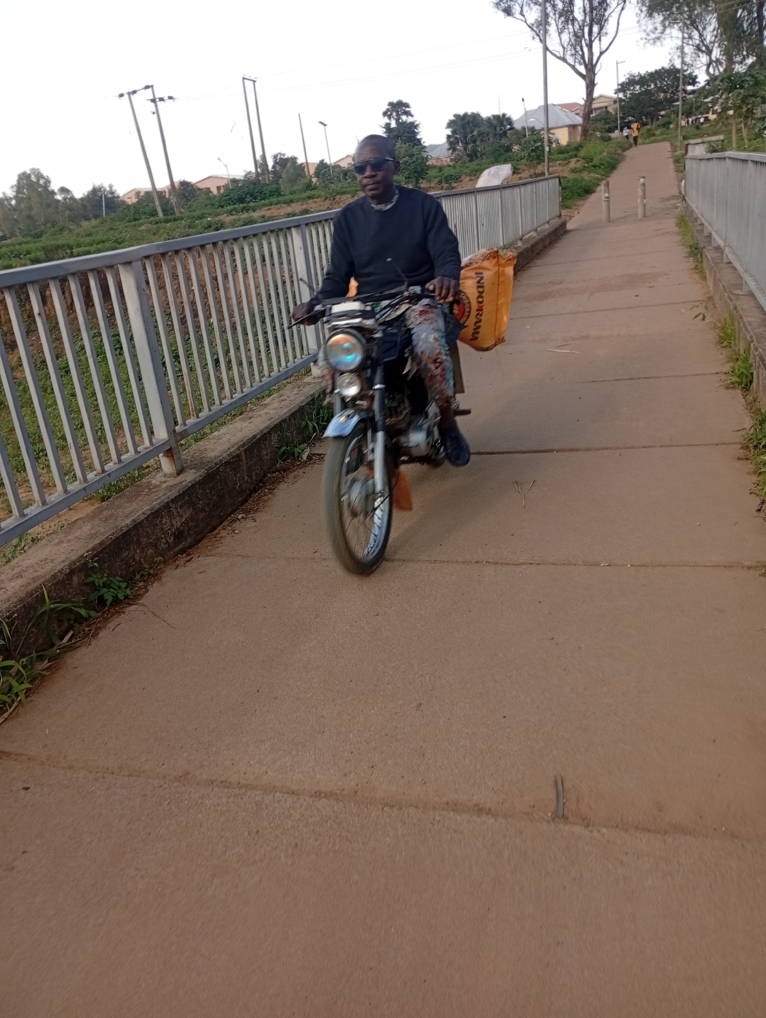 A person rides a motorcycle on a paved path. The person is wearing sunglasses and a dark long-sleeved shirt. They are carrying a large orange bag on the back of the motorcycle. The path appears to be a pedestrian bridge with a metal railing on the left. In the background, there are trees, buildings, and power lines, suggesting an urban or semi-urban environment. The overall impression is a daytime scene, with natural light illuminating the area. The location is specified as Rafin Par, Nigeria.