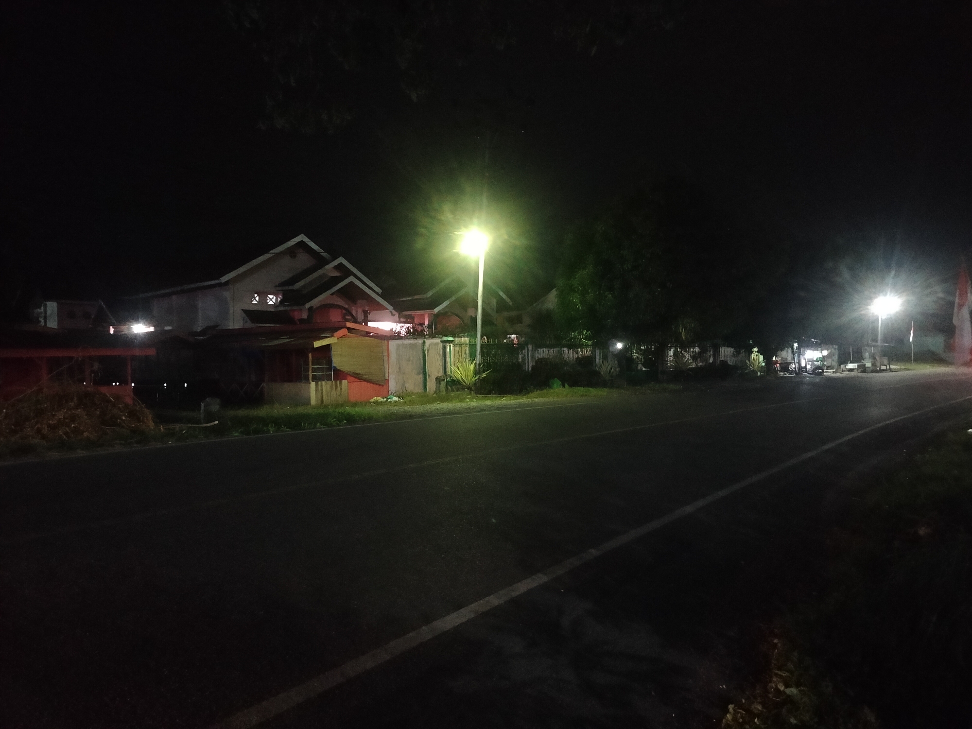 A nighttime street scene in Kumbangjaya, Indonesia, features a dark asphalt road with a white line marking the lane. On the left side of the road, a low-rise building with a red trim and a light-colored facade is visible, with a pile of dried vegetation in front. Further down the road, a bright streetlamp illuminates the area. Beyond the building, more structures and trees are dimly lit. On the right side of the road, the landscape is dark with some scattered lights and vegetation. The overall atmosphere is dark and quiet, typical of a rural or suburban area at night.