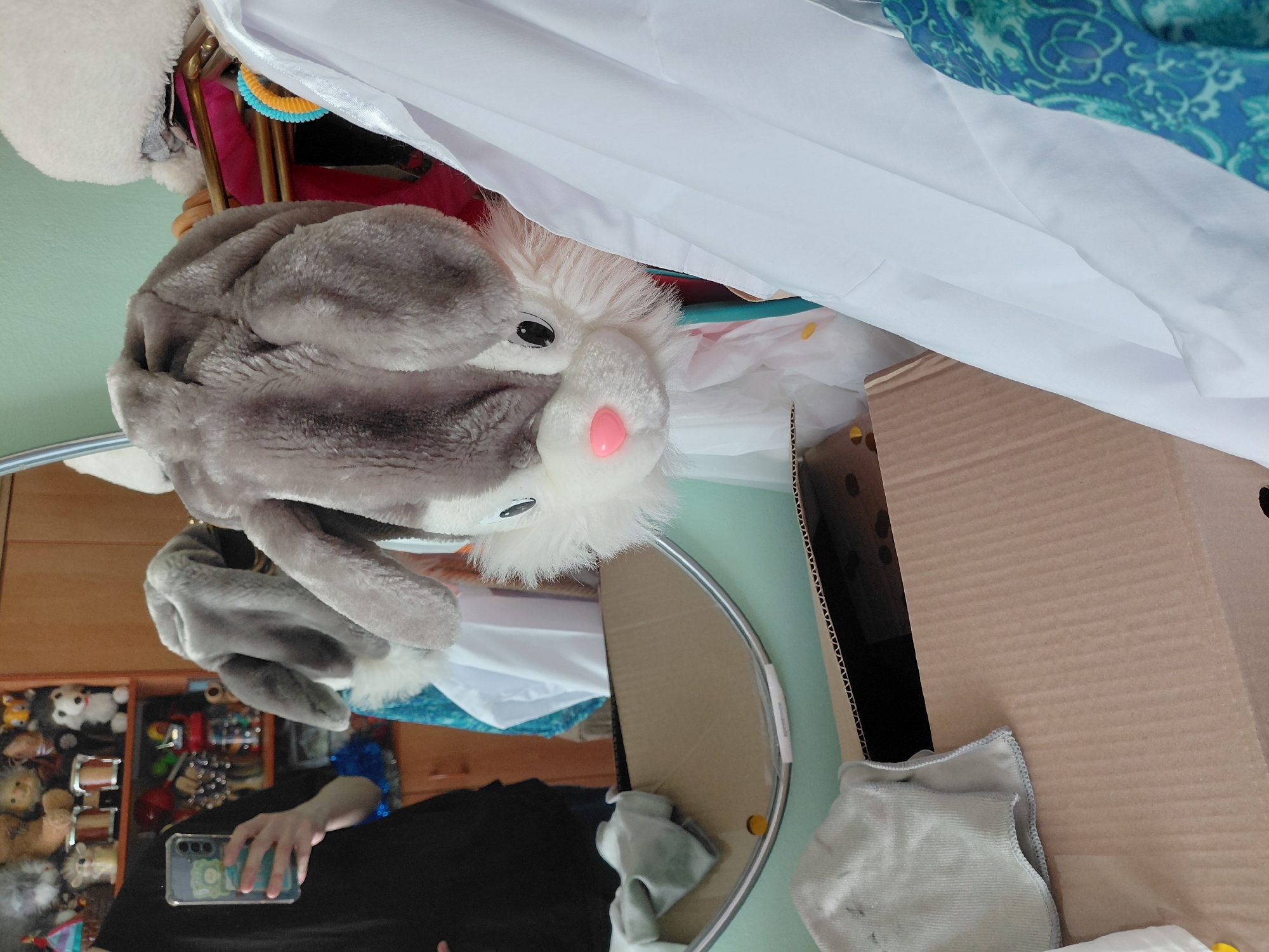 An indoor, vertically oriented mirror selfie captures a cluttered domestic setting. The main subject is a large, gray and white plush rabbit or bunny, prominently featured in the upper-center of the frame, with a fluffy white face and a pink nose. Below it, a person's hand, wearing a black sleeve and a blue cuff on the wrist, holds a light-colored smartphone with a graphic design on its case, positioned to capture the reflection.
The oval mirror, through which the photo is taken, reflects a wooden cabinet or shelf unit in the background, filled with numerous stuffed animals, toys, and various other household items. To the right of the mirror, in the actual room, a large, open cardboard box is visible. A light green wall forms part of the background. Above the plush toy, white and patterned blue fabrics or clothing hang, alongside a small yellow and orange decorative item. A white fluffy object is visible in the top left corner, possibly another plush toy or a hat. A gray cleaning cloth rests on a surface near the cardboard box. The setting suggests a personal space, possibly a bedroom or playroom. No discernible text is visible on any objects. The activity is a person taking a mirror selfie with a plush toy. The location context provided is Nadym, Russia.