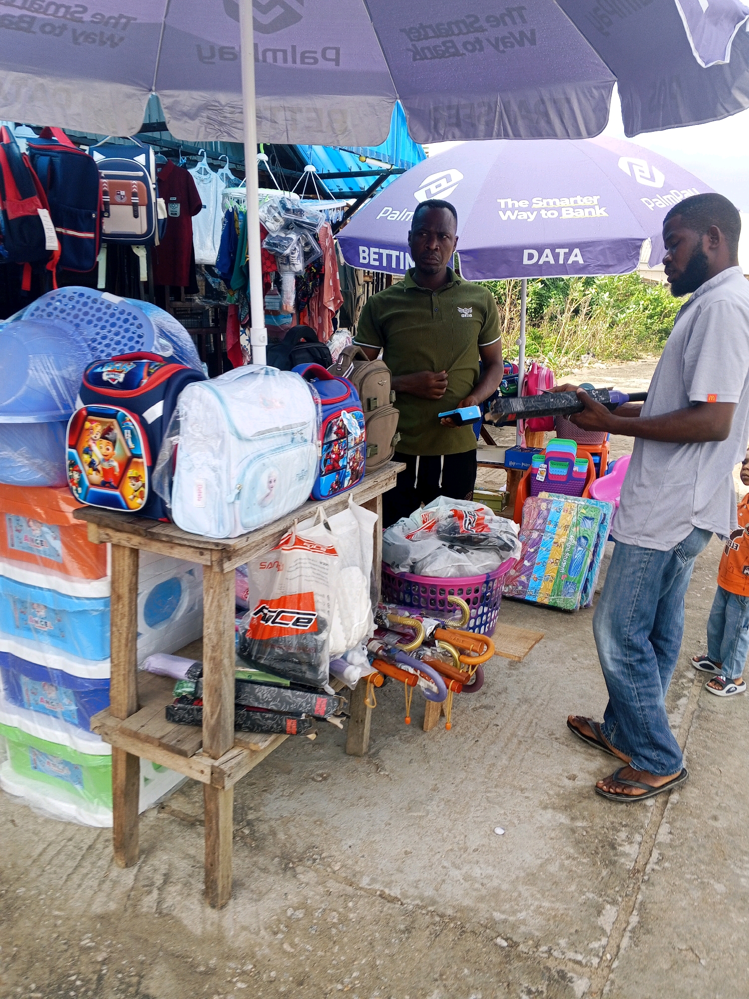 In Zariagi, Nigeria, two men and a child are gathered at an outdoor stall under large purple umbrellas. The stall is laden with various goods, including backpacks, plastic storage containers, and folded items wrapped in plastic. One man, wearing a green polo shirt, stands behind the stall, while the other, in a grey t-shirt and blue jeans, faces him. The man in grey is holding an item wrapped in black plastic. A child in an orange shirt stands to the right. The ground is paved with concrete, and the background shows vegetation.