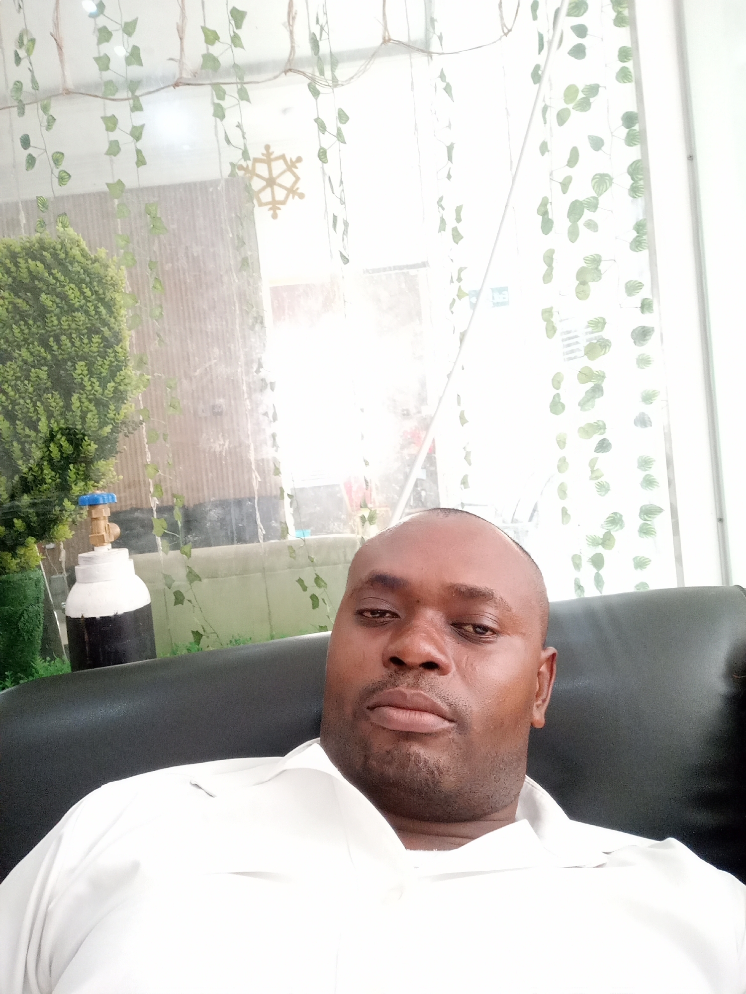 A man is featured in a close-up selfie, seated indoors in what appears to be a dark chair or sofa. He has dark skin, a shaved head, and is wearing a white collared shirt. His expression is neutral as he looks directly into the camera.
The background is brightly lit, primarily through a large window or glass partition. Artificial green vines with small leaves are decoratively strung across the top and hang down within this transparent barrier. A golden, snowflake-shaped ornament is also suspended in the upper background.
To the left of the man, behind the glass, is a lush green potted plant or shrub. Adjacent to the plant stands a white and black cylindrical object topped with a blue and brass valve, resembling a small oxygen tank. Beyond the partition, glimpses of a light-colored corrugated structure and a light green surface suggest an exterior or an adjacent bright space.
The lighting is bright, indicative of daytime. The setting appears to be a casual indoor environment, possibly a waiting area, office, or lounge in Gwarinpa, Nigeria. There is no visible text in the image.