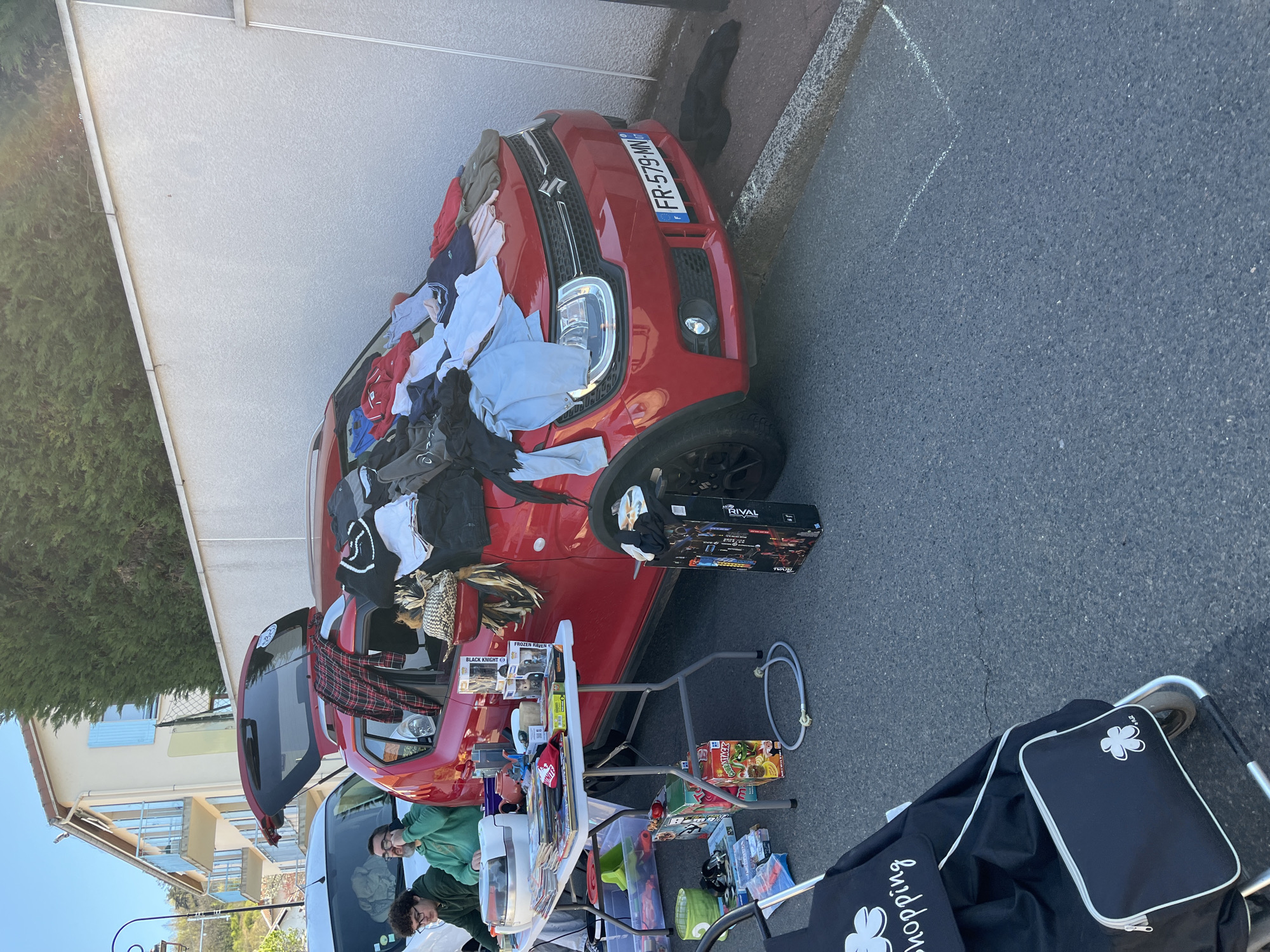 The image shows a flea market setup next to a red Suzuki Ignis car (license plate FR-579-MN). The car has clothes draped on the hood. Items for sale are displayed on a folding table, including games, books, figurines, and other miscellaneous objects. Two people are visible behind the table. A black shopping cart is positioned to the right of the table. The location appears to be a residential street, as a building is visible in the background. Based on the visual context, this event is likely taking place in Bellegarde-en-Forez, France, as the license plate is from France. The scene depicts a typical outdoor flea market setting.