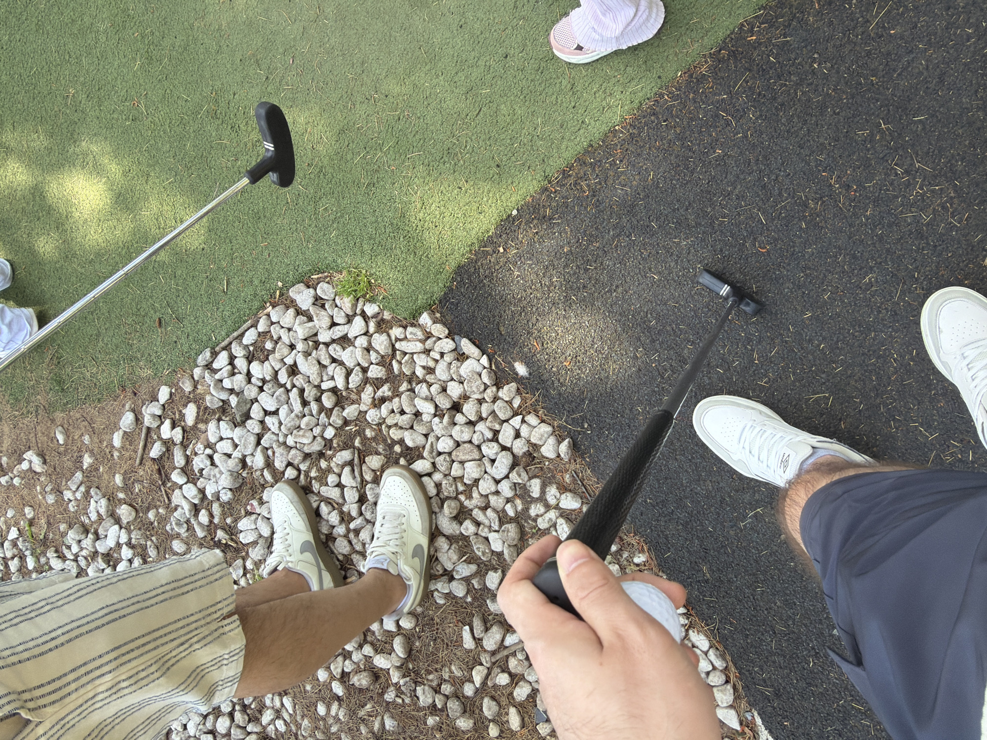 The image shows a view from above of four people playing mini golf. In the foreground, a hand holds a golf ball and putter. The person is wearing navy shorts and white sneakers. To the left, another person is wearing striped shorts and white sneakers, standing on small white rocks. To the left of that person, a golf putter lies on a green artificial turf surface. To the upper right, another person is wearing white shoes and is standing on a black artificial turf surface. A fourth person is present in the upper left corner of the image wearing pink sneakers and is also standing on the green artificial turf surface. The ground is divided into sections of artificial turf, small white rocks, and black artificial turf. The location is likely a mini golf course in Warrington, United Kingdom.