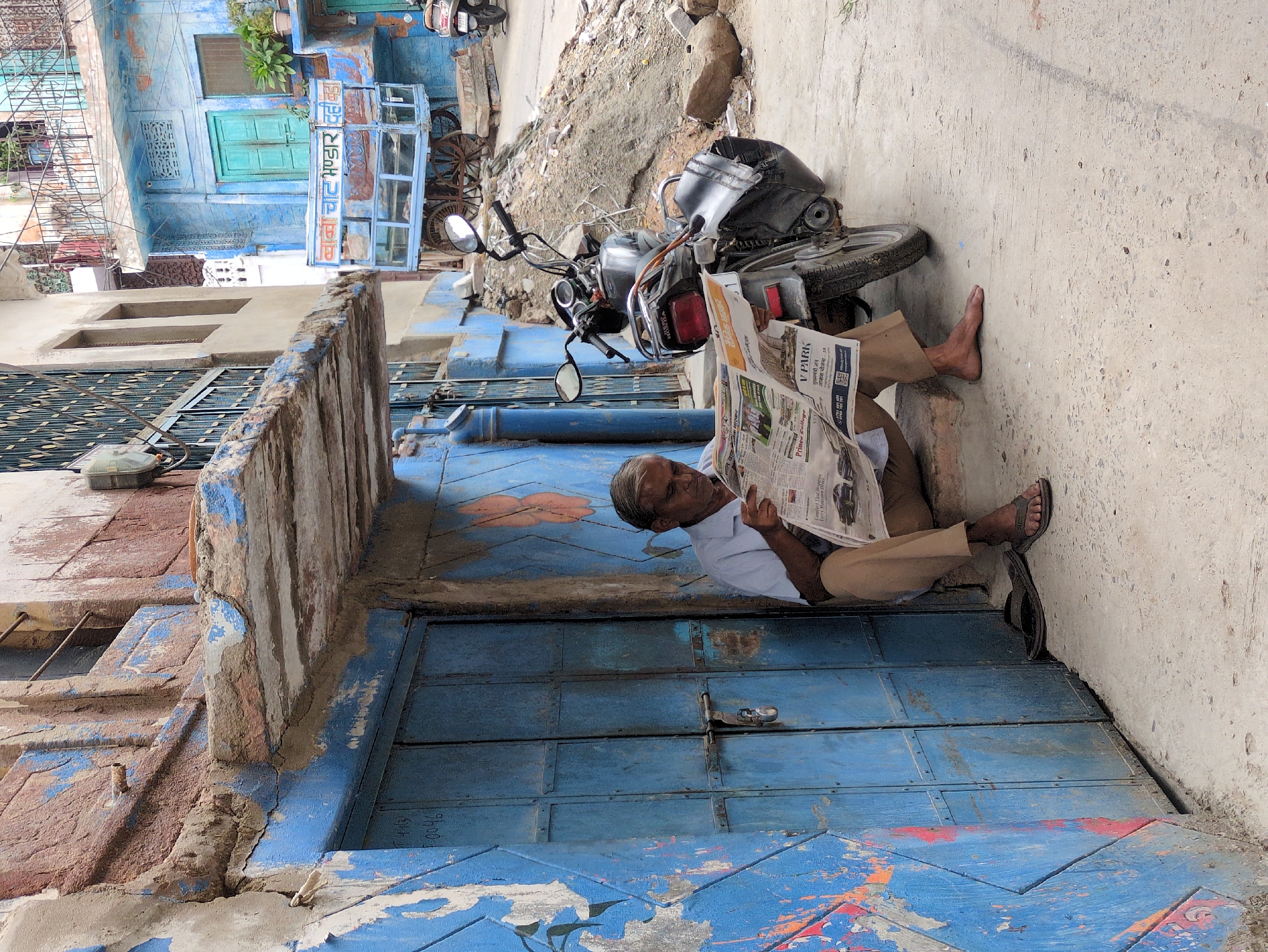 A man sits on a low step against a weathered blue door, reading a newspaper. He is wearing a light blue collared shirt and tan pants. His left foot is bare, and his right foot is clad in a dark sandal. A motorcycle is parked to his right, leaning on its kickstand. The building behind the man and the motorcycle has a textured, peeling facade with blue paint on the door and surrounding walls, and a reddish-brown upper section. In the background, a blue cart with a glass display case and large wheels is visible, as is another building painted blue with green doors. The ground is a rough, textured concrete. The scene appears to be in a narrow street or alleyway in Jodhpur, India, characterized by its blue architecture and vibrant, worn textures.