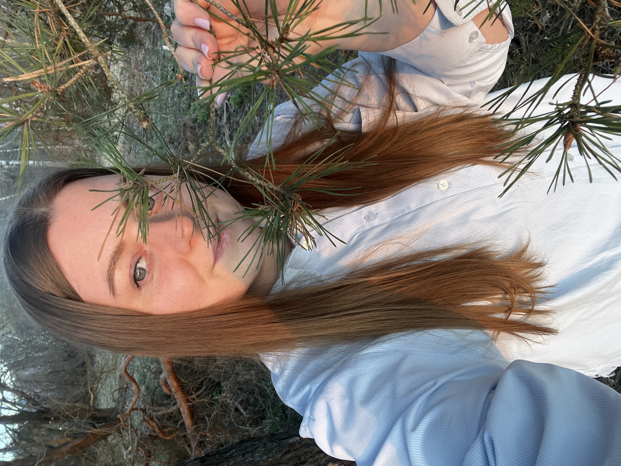A light-skinned woman with light brown hair is pictured from a high angle. She has one visible blue-green eye and wears a light blue, collared shirt. A pine branch partially obscures her face and she holds the branch with her hand. The background appears to be woodland with visible tree trunks and ground vegetation. The exact location in Voronino, Russia, is not determinable from the image.
