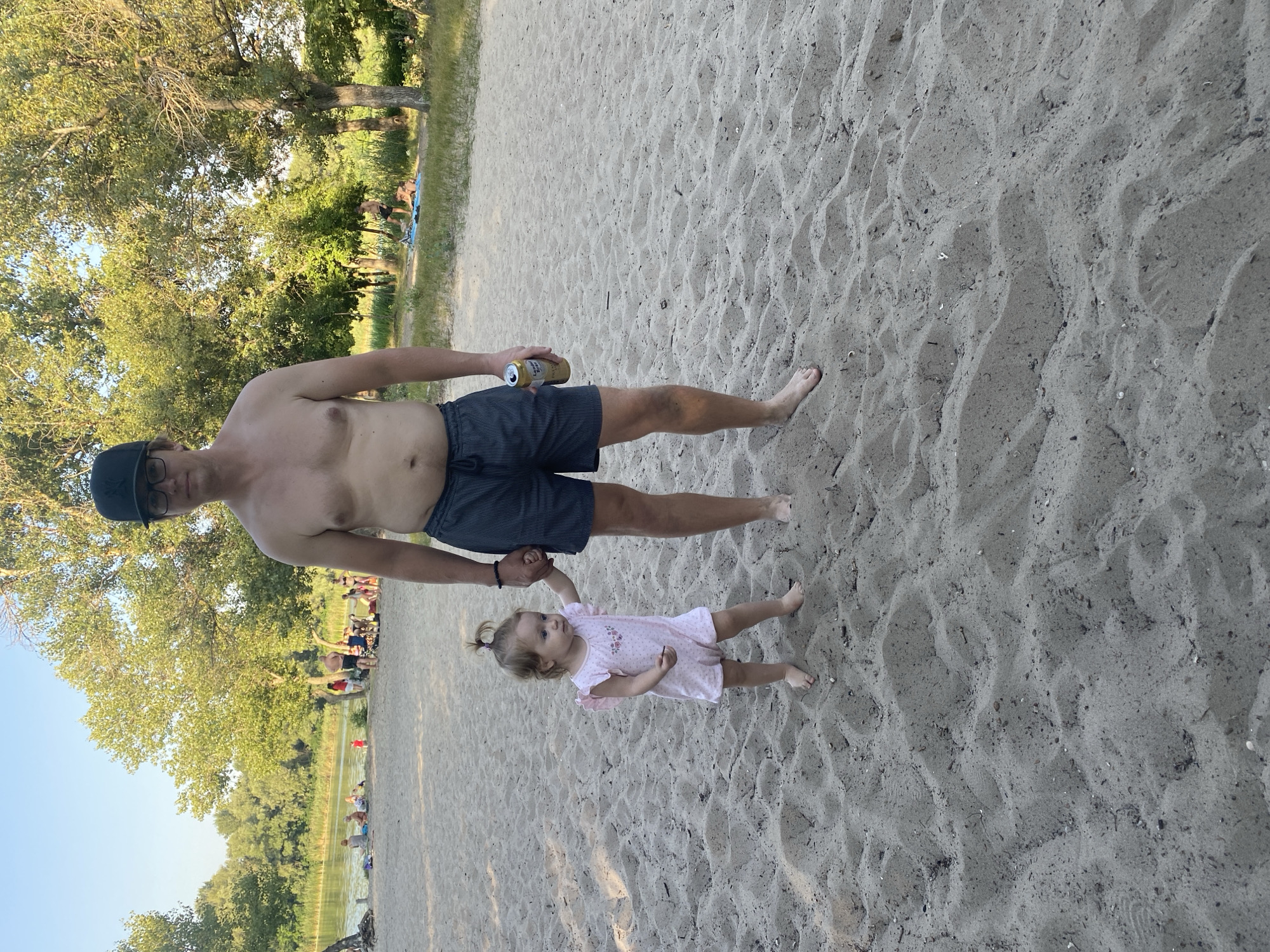A man and a young child stand on a sandy beach. The man is shirtless and wearing dark shorts and a baseball cap. He is holding the child's hand. The child is wearing a light-colored romper with a floral pattern and has a ponytail. The man is holding a can in his right hand. In the background, there is a body of water, trees, and other people on the beach. This scene likely depicts a day at the beach on the Dnipro River in Dnipro, Ukraine.